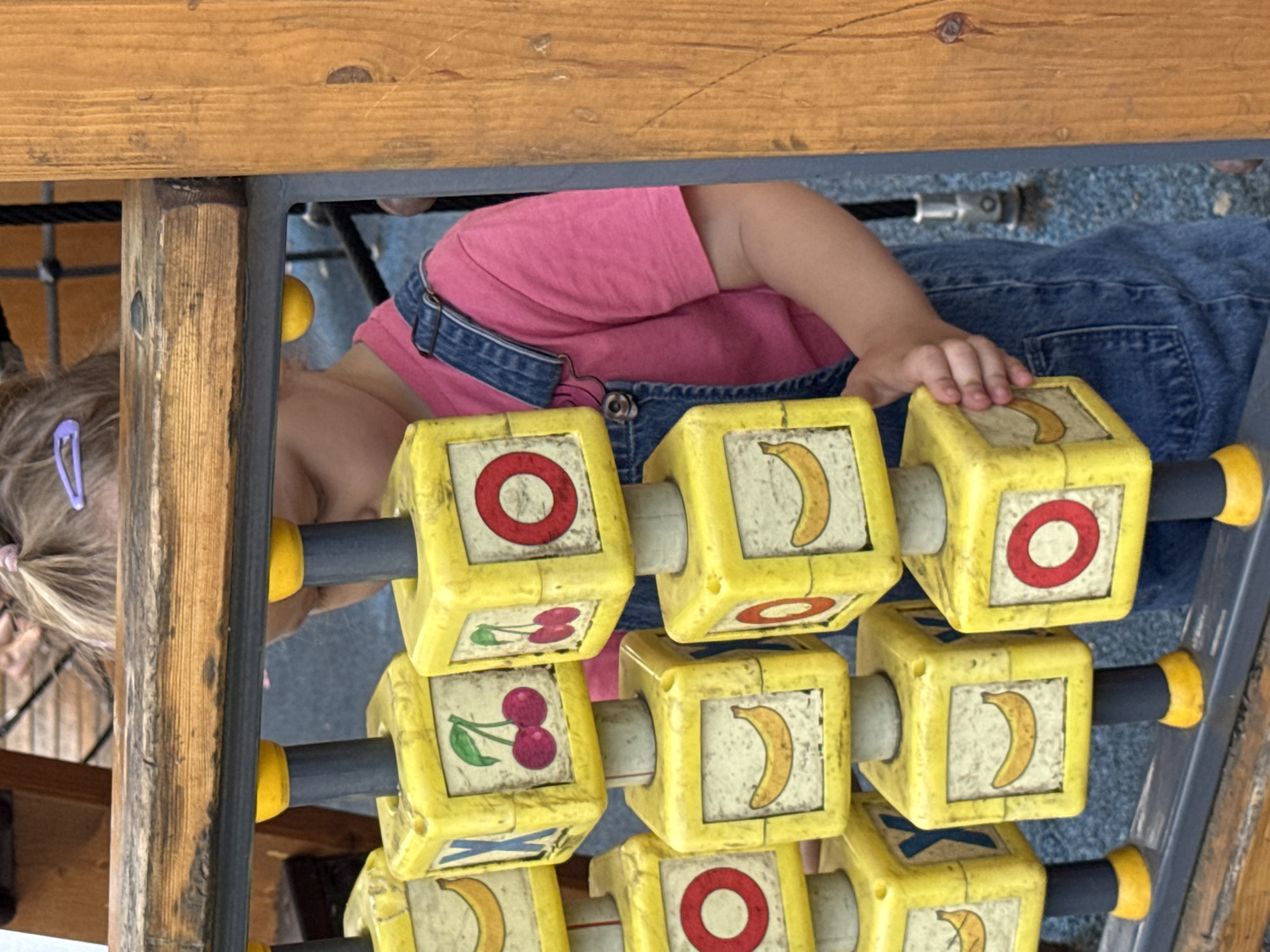 A young child, wearing a pink t-shirt and denim overalls, is playing with a wooden toy made of yellow blocks. The blocks are square and have images of bananas, cherries, and red circles printed on them. The toy is part of a playground structure. The child's face is partially obscured by the toy. The background shows wooden planks and a blue surface that appears to be the ground of the playground. The location is the city of Mikhaylovsk, Russia.