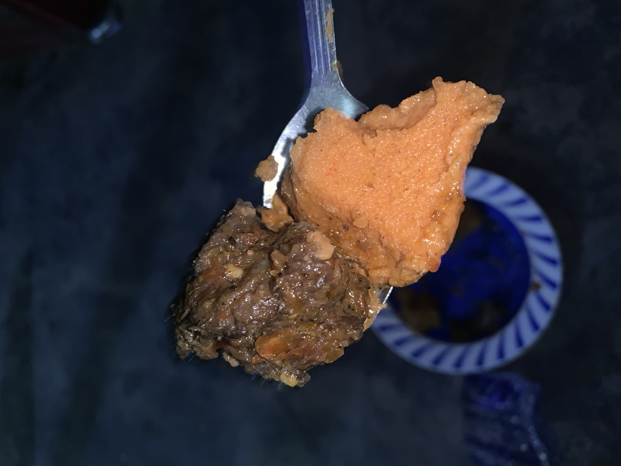 A spoonful of food, consisting of two distinct components, is held aloft. The upper portion is an orange, pasty substance. Below it is a dark brown, chunkier mass with visible fragments of carrots. A blue and white striped bowl is in the blurred background. The location is the city of New Stadium, Nigeria.