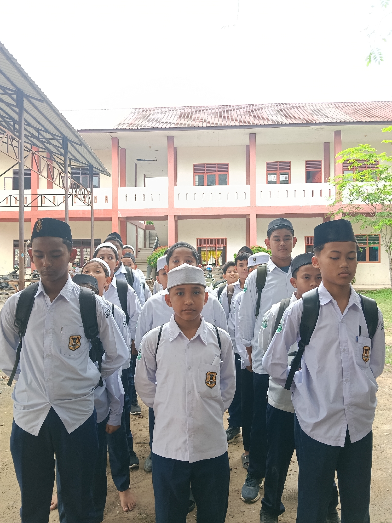 A group of young male students stand in rows outdoors in what appears to be a schoolyard. They are uniformly dressed in white shirts and dark-colored pants, with some wearing backpacks. Some of the boys wear traditional Muslim caps (peci or kopiah) on their heads. They are lined up in a formal arrangement. The background includes a two-story building with a red tile roof and light-colored walls. A metal-framed structure, possibly part of a covered walkway, is visible on the left. The environment suggests a school setting in Lhokseumawe, Indonesia.