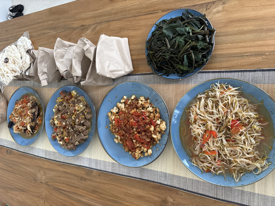 The image displays a meal laid out on a light-colored wooden table, suggesting an indoor setting in Canggu, Indonesia. A blue and white striped fabric table runner is positioned vertically along the table's length.
Five blue ceramic plates hold various dishes. Four larger blue plates are arranged in a vertical column on the table runner:
1. The top plate contains a dark-colored meat or seafood dish topped with a generous amount of finely chopped red chilies and light-colored aromatics, consistent with sambal matah.
2. The second plate from the top features diced, cooked meat mixed with scrambled egg pieces, red chili fragments, and other seasonings.
3. The third plate from the top presents diced white cubes, consistent with tofu or tempeh, heavily garnished with fried shallots and sliced red chilies.
4. The bottom plate displays stir-fried bean sprouts with visible slices of red chili and tomato in a light, clear sauce.
To the right of the third and fourth plates, a smaller blue plate contains a mound of dark green, cooked leafy vegetables, resembling water spinach or similar greens.
Adjacent to the right side of the main column of plates, five individual portions, each wrapped in a light brown paper bag, are arranged vertically. A transparent plastic bag filled with numerous white, irregular-shaped crackers, bearing a small black circular sticker, rests on the uppermost brown paper bag.
In the upper right corner of the frame, two black flip-flop sandals are partially visible on a light-colored floor. No individuals are present, and no actions or interactions are depicted.