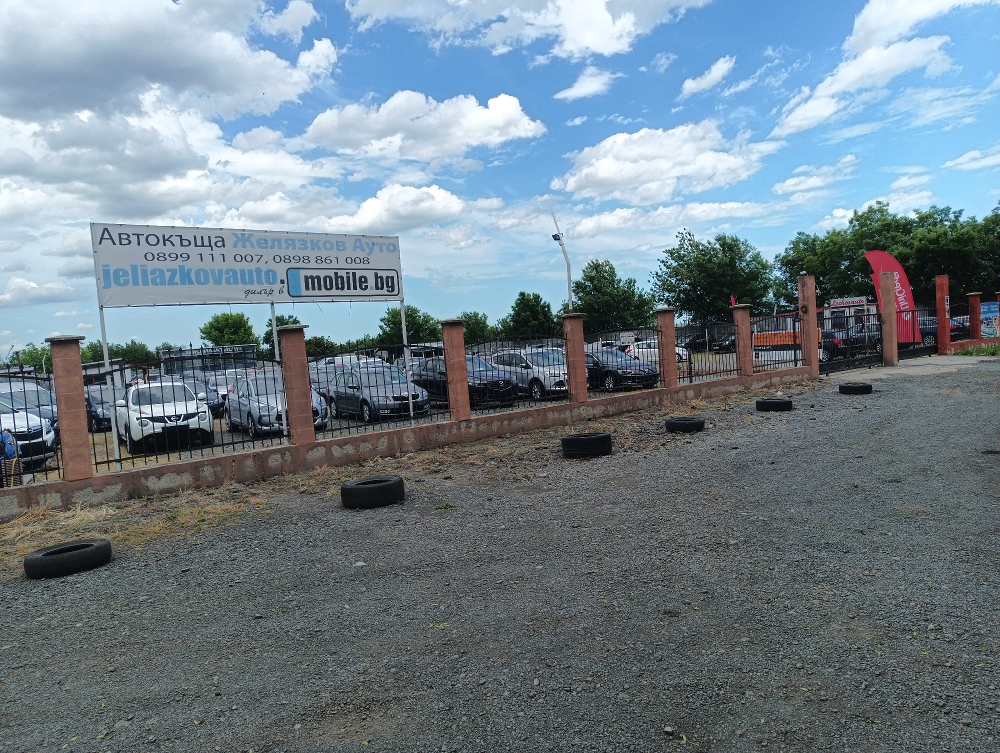 The image shows an outdoor car lot in Burgas, Bulgaria, presumably "Автокъща Желязков Ауто" (Zhelyazkov Auto). A sign bearing the company name and phone numbers (0899 111 007, 0898 861 008) is prominently displayed, along with the website "jeliazkovauto.mobile.bg". A variety of cars are visible behind a low fence made of brick pillars and black metal bars. The ground is covered in gravel and scattered with used tires. The sky is partly cloudy. A red flag is visible in the background.