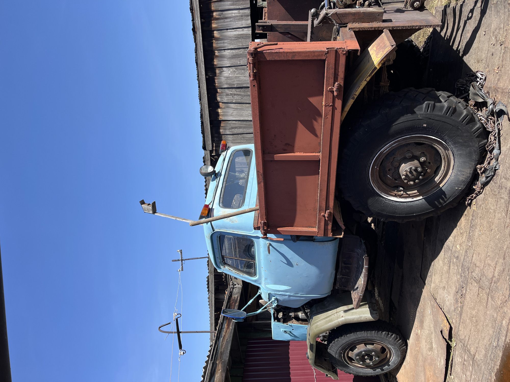 A faded blue and red dump truck sits adjacent to a weathered wooden building with a rusty corrugated metal lower section in Верхняя Уря, Russia. The truck's tires are thick and worn, with one tire wrapped in chains. The sky above is clear blue. Utility poles with wires are also visible. The ground appears to be covered with wooden planks. The truck appears old and in a state of disrepair.