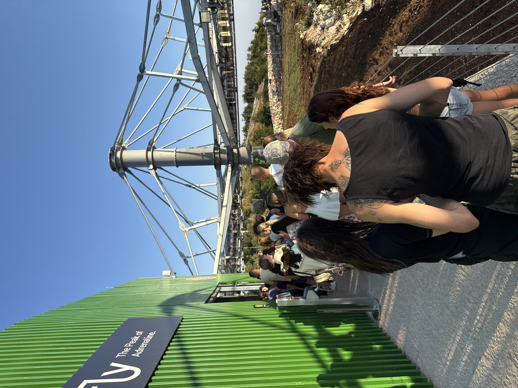 In the city of Kuk, Montenegro, a group of people are queuing outside a green building that has a sign reading "The Peak of Adrenaline." The building's facade is constructed from vertical corrugated metal panels. A metal structure with cables, possibly part of an attraction, extends into the background. The ground is paved with light-colored stones. The sky is clear and blue. The people in the queue are diverse in age and attire, with some facing away from the camera and others looking towards the entrance.