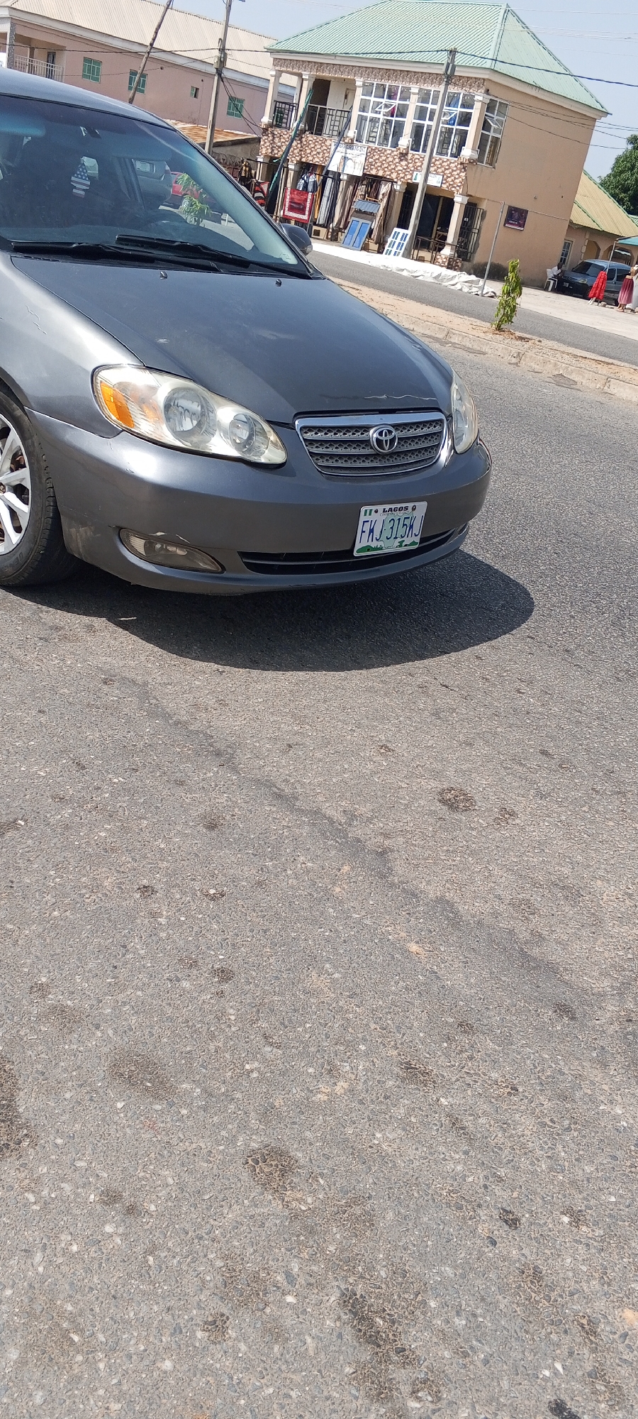 A gray Toyota Corolla is parked on a paved road in Jalingo, Nigeria. The car's license plate reads "FKJ 315KJ" and indicates "LAGOS". In the background, a two-story building with a green roof houses shops on the ground floor and apartments above. The shops display textiles, rugs, and windows. The street is bordered by a curb and sidewalk. Sunlight casts shadows on the road, indicating it is daytime.