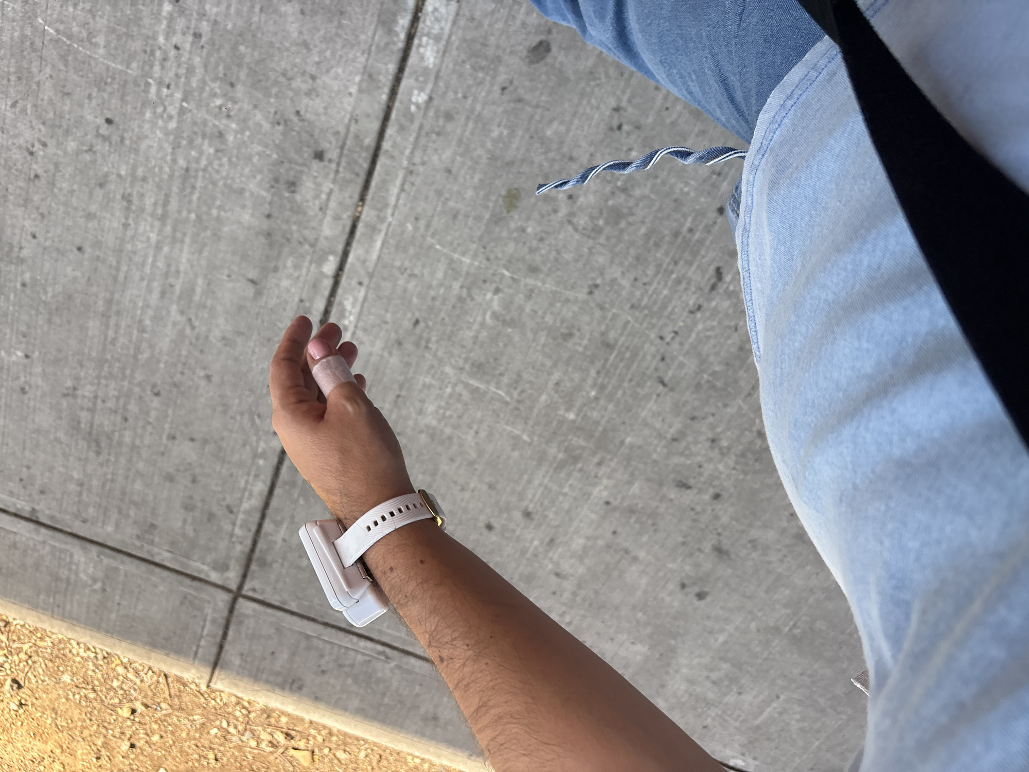 An overhead shot captures a person outdoors, focusing on their arm, hand, and a portion of their leg. The person's left arm extends towards the bottom left of the frame, revealing a light-skinned forearm with some hair. A white wristband, possibly a smartwatch or fitness tracker, is worn on the wrist, featuring a gold-colored buckle. Attached to the underside of the wristband is a larger, rectangular white device. The hand shows a bandage wrapped around one finger, likely the ring or pinky finger.
The person's upper thigh or hip area is visible on the right, clad in light blue denim fabric, possibly jeans or shorts. A twisted blue and white drawstring or cord hangs from this garment. Part of a black strap, possibly from a bag or belt, is also visible.
The setting is an outdoor concrete sidewalk or pathway, characterized by a grey, textured surface with visible cracks and lines. To the lower left, a patch of dry, light brown soil or gravel with small stones can be seen, bordering the concrete. The bright lighting and clear shadows indicate a sunny daytime environment. No specific activity beyond standing or walking is clearly depicted, and no text is visible. The scene suggests a casual moment in an urban or suburban area, consistent with Mexico City.