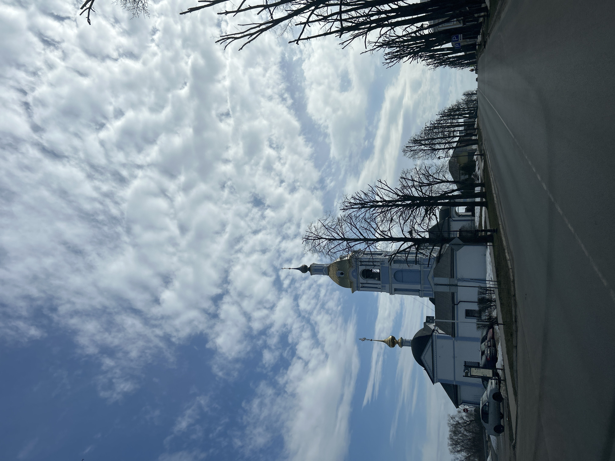 The image depicts a street scene in Unknown, Unknown. A road runs along the right side of the frame. Bare trees line the street and a church is visible on the left. The church has white walls and gold domes. A blue sky filled with clouds stretches above the buildings and trees. Several cars are parked beside the church.
