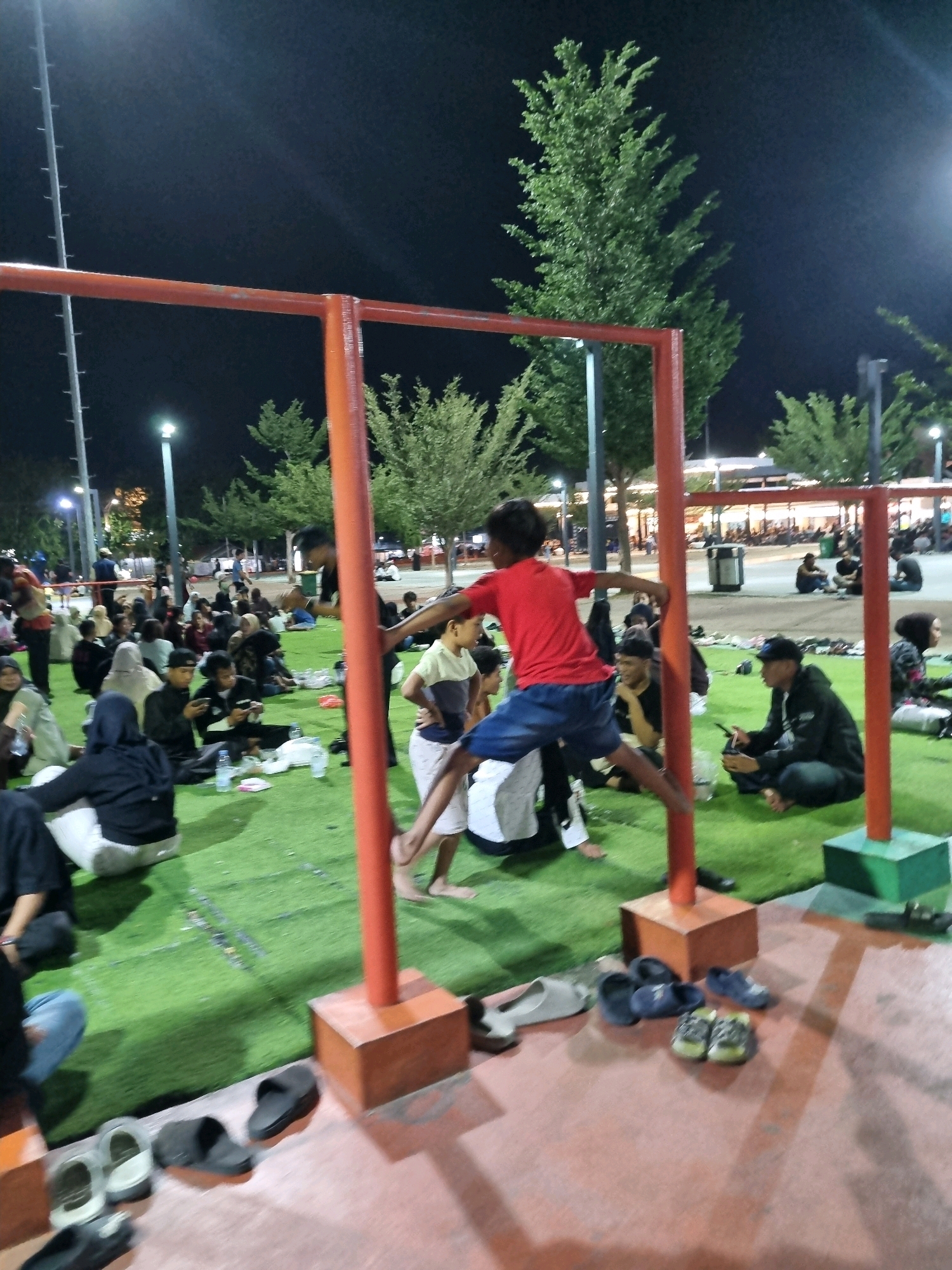 In Tanamodindi, Indonesia, a night scene unfolds in an outdoor recreational area. A group of people, appearing to be youth, are gathered on a vibrant green artificial turf. A young male, wearing a red t-shirt and blue shorts, is actively engaged in a physical activity, positioned mid-jump or swing on a red metal bar structure. This structure, consisting of horizontal and vertical bars, is set up on the turf. The background reveals a dimly lit environment with trees and artificial lighting. Other individuals are seated and standing around the turf, some looking towards the boy on the bars, while others are engaged in their own activities, with some holding mobile phones. Several pairs of shoes are scattered on the ground in the foreground.