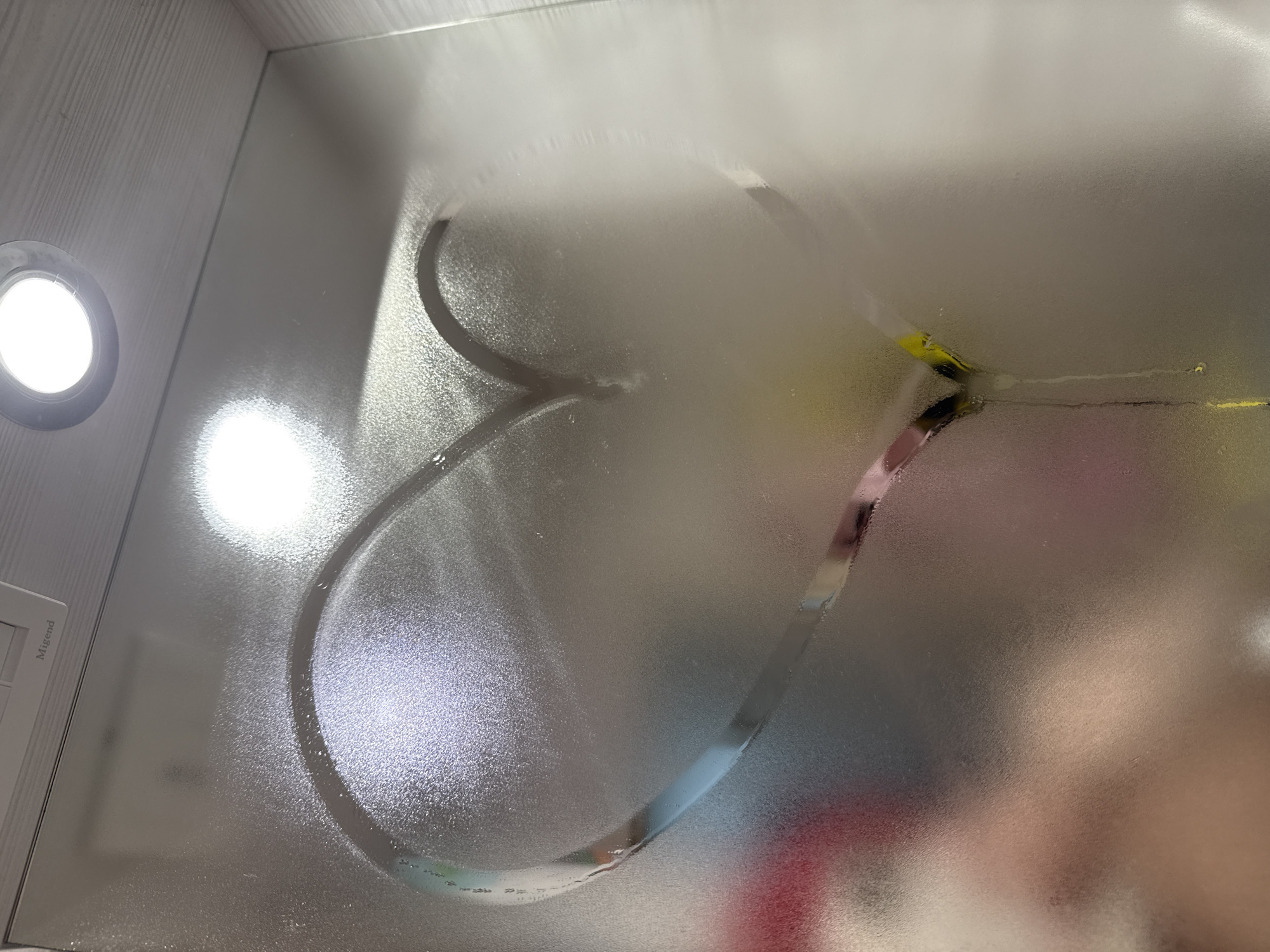 The image displays a heart shape drawn onto a heavily condensed or steamy glass surface, likely a mirror or shower door. The heart has been wiped clear, removing the fog and allowing a view of the reflections and objects behind it. Through the cleared outline of the heart, blurred colors such as yellow, pink, blue, and red are visible, suggesting colorful items or reflections in the background.
On the left side of the frame, a bright, circular light fixture with a strong white glow is prominently featured. Another, softer circular light is faintly visible in the upper center of the image. To the far left, a white rectangular panel, which appears to be a light switch or control unit, has the word "Migend" printed on its surface.
The activity depicted is the act of drawing a heart on a steamy surface, conveying a sense of affection or playfulness. The setting is an indoor environment, strongly suggested to be a bathroom due to the condensation and light fixtures. The visible text "Migend" identifies a brand or label on a fixture. The overall scene captures a momentary, intimate gesture in a common domestic setting.