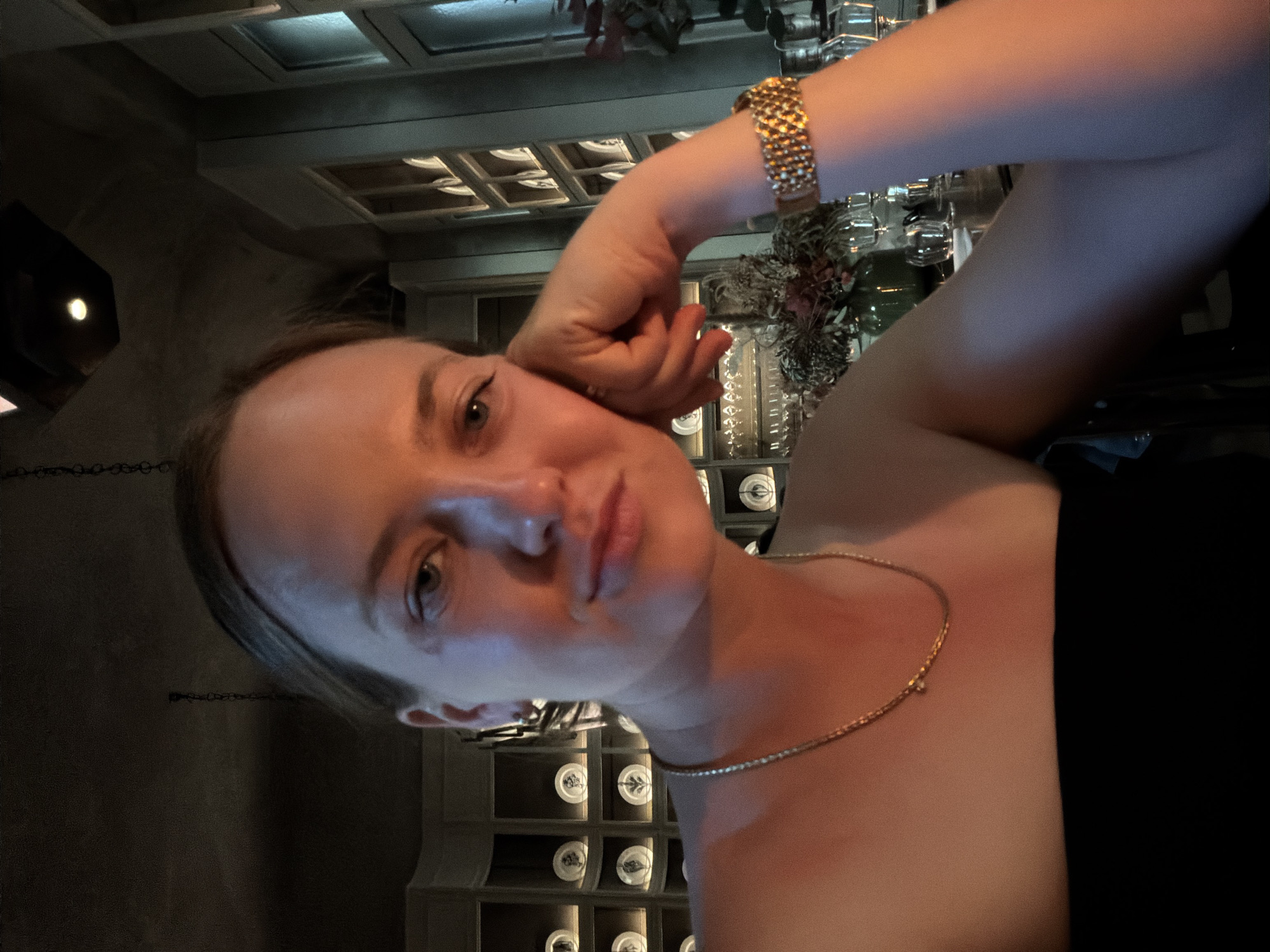 A woman, with blonde hair pulled back, is captured in a close-up selfie. She is leaning her head on her hand, her gaze directed towards the camera with a slight smile. She is wearing a black strapless top and a delicate gold chain necklace. A gold bracelet adorns her wrist. The background suggests an indoor setting with warm, ambient lighting. Shelves lined with decorative plates are visible behind her. The overall atmosphere is intimate and dimly lit, possibly in a restaurant or a stylish lounge. The location is identified as Milan, Italy.