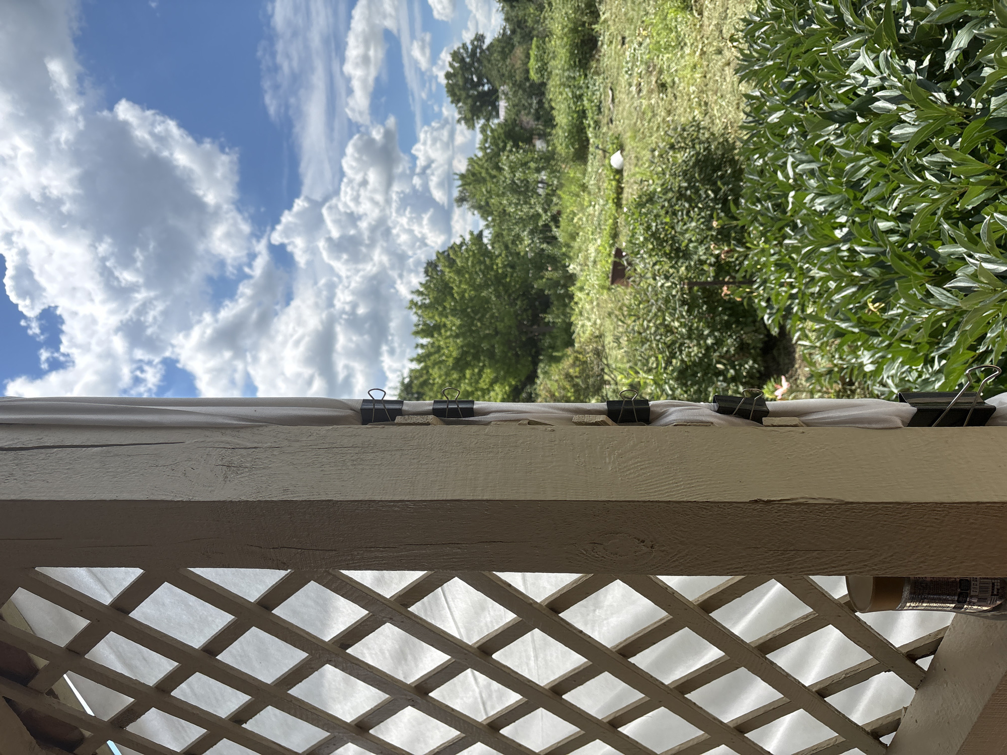 The image depicts an outdoor scene with a clear blue sky filled with fluffy white clouds, suggesting it is daytime and the weather is fair. The perspective is from under a wooden structure, possibly a porch or gazebo, with a lattice pattern visible in the foreground. This lattice is made of light-colored wood and has a diamond-shaped design, allowing a view of a white, translucent material behind it.
Above the lattice, a horizontal wooden beam runs across the image. This beam is painted a light, creamy color and shows signs of wear, with some grain visible. Along the top edge of this beam, a white material is held in place by several black binder clips, also secured with small pieces of wood.
Beyond the wooden beam, the scene opens up to a lush green landscape. Trees with full green foliage dominate the mid-ground, interspersed with shorter bushes and grassy areas. The greenery is vibrant, indicating it's likely springtime or summer. In the far distance, more trees can be seen against the sky. The overall impression is one of a natural, possibly semi-rural or suburban setting on a pleasant day. There is no visible text in the image.