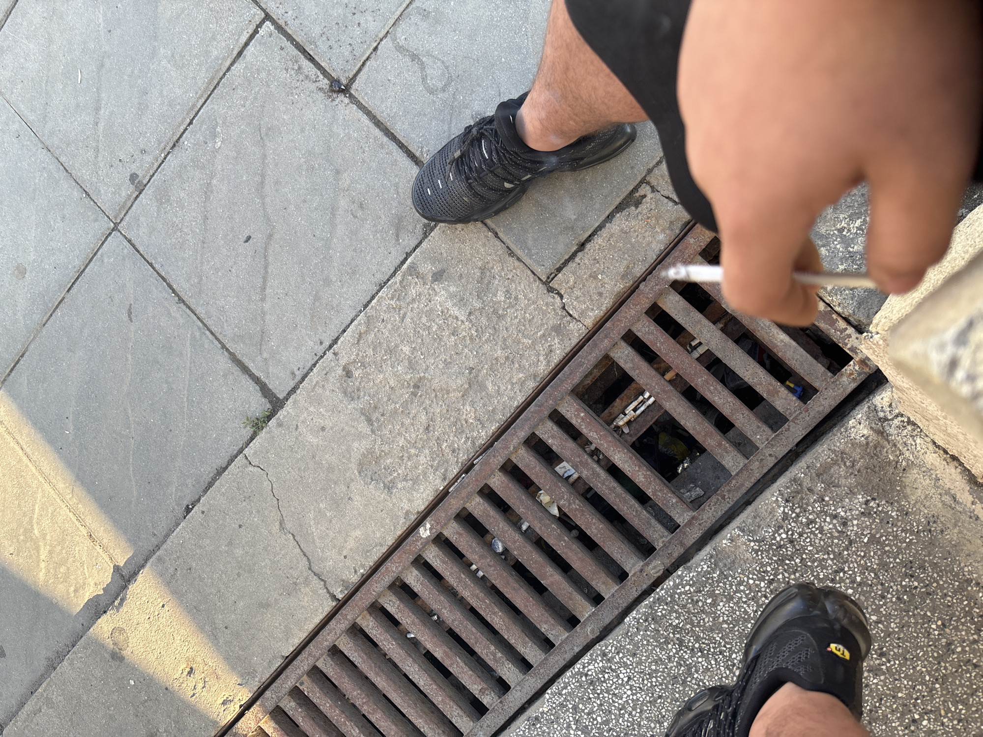 An overhead, high-angle shot captures a person standing on a paved surface next to a storm drain. The scene primarily features the person's legs, feet, and right arm/hand, along with the ground beneath them.
The person is wearing black shorts and black athletic sneakers. Their right leg is partially visible on the left side of the frame, and their left leg and foot are in the bottom right corner. The right hand holds a lit cigarette, which is positioned directly over the metal storm drain grate. The fingers of the hand are slightly blurred.
The ground consists of grey concrete pavement. On the left side, it's laid in large square tiles. A distinct, sharp diagonal shadow from an unseen object or building cuts across the lower-left section of the pavement, indicating strong sunlight. Towards the right side and around the drain, the pavement appears to have a rougher, possibly gravel-like texture.
The storm drain is a long, rectangular metal grate running diagonally across the middle of the frame. Through the grates, various pieces of litter are visible, including several discarded cigarette butts and some plastic debris.
On the toe of the left black sneaker, a small yellow logo with the text "Tn" is visible. The overall setting appears to be an urban or suburban outdoor environment during daytime with clear weather.