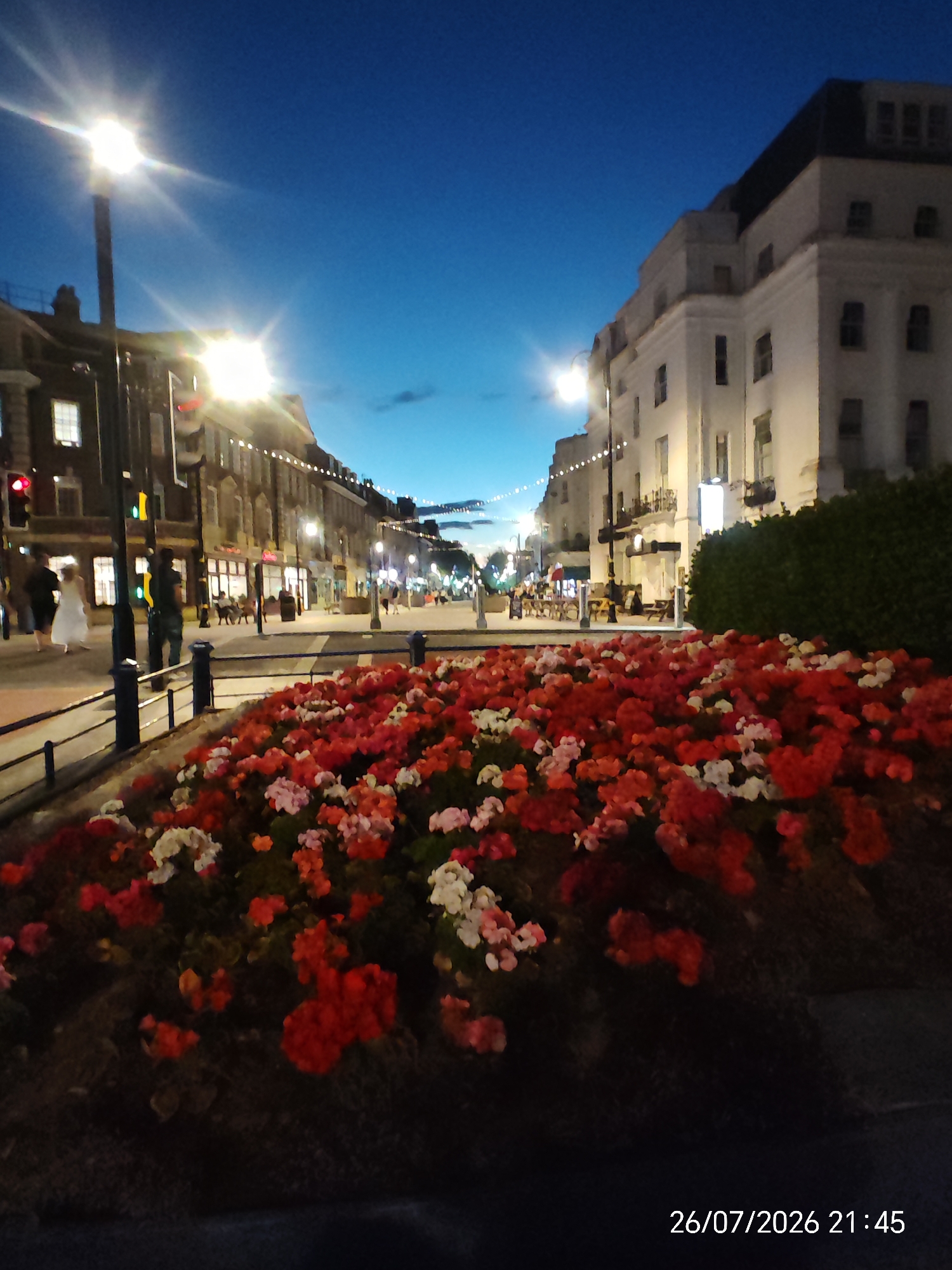 A nighttime street scene in Eastbourne, United Kingdom, features a prominent flowerbed in the foreground filled with red, pink, and white flowers. Behind the flowerbed, a street lined with buildings and streetlights extends into the distance under a dark blue sky. People are visible walking on the sidewalk and in the street. One person in a white dress is walking with another individual. Streetlights illuminate the scene, and a traffic light is visible. The architecture suggests a commercial or residential area with multi-story buildings. A timestamp, "26/07/2026 21:45," is present in the bottom right corner.