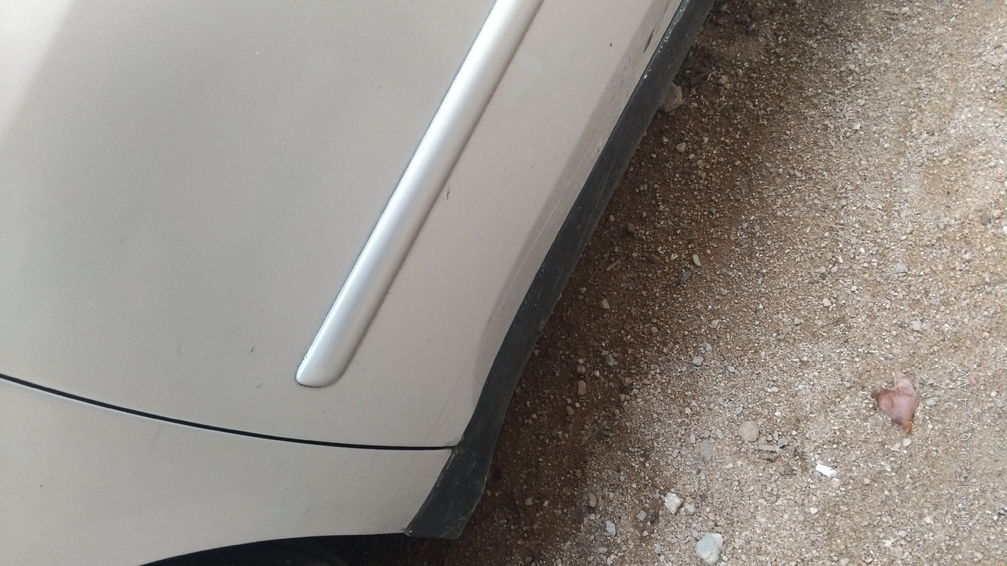 This is a close-up, eye-level shot of the lower portion of a light-colored car door and the sandy ground beneath it. The car appears to be parked on a dirt or gravel surface, possibly a roadside or an unpaved area. A black plastic trim piece runs along the bottom edge of the car door, partially obscured by the ground. A subtle chrome or silver trim piece is also visible higher up on the door. The ground is composed of small stones, pebbles, and dirt. There are a few small, scattered objects on the ground, including a light-colored piece of debris and a reddish-brown organic item. The lighting suggests it is daytime. There is a watermark in the bottom left corner that reads "June 15, 2026 7:24 AM". The scene is devoid of any human presence. The location in Howayi, Nigeria cannot be determined from the visual information provided.