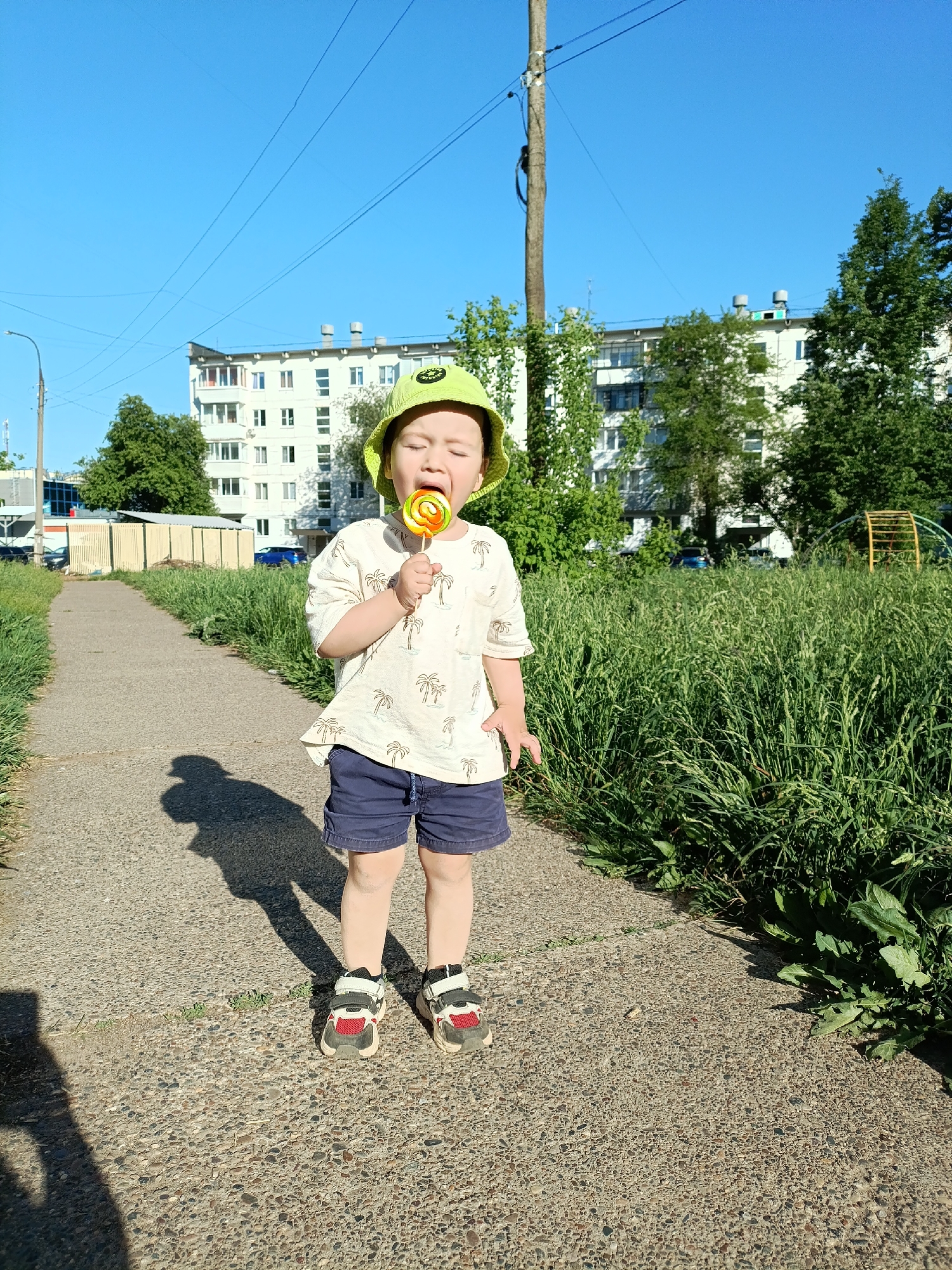 A young child stands on a concrete pathway, sucking on a colorful lollipop. The child wears a light-colored T-shirt with a palm tree pattern, navy blue shorts, and sneakers with red and white accents. A light green bucket hat sits on the child's head. The child's eyes are closed as they enjoy the lollipop.
The pathway is flanked by patches of grass and weeds. In the background, a multi-story white apartment building is visible, along with trees and a wooden utility pole. A partial view of a playground structure can be seen to the right of the building. The scene appears to be in a residential area, likely in the city of Chaikovsky, Russia. The lighting indicates it is daytime.
