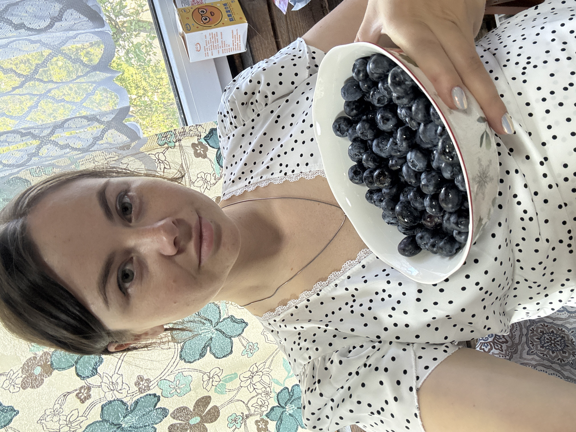 A woman is holding a white bowl filled with blueberries. The bowl is tilted towards the camera. She is wearing a white top with black polka dots and lace trim. A thin necklace is visible on her neck. Her fingernails are painted with a light, reflective polish. In the background, there is a window with a patterned curtain, a partially visible box with Chinese characters, and an outdoor scene. The location appears to be in the city of Малая Рукавицкая, Russia.