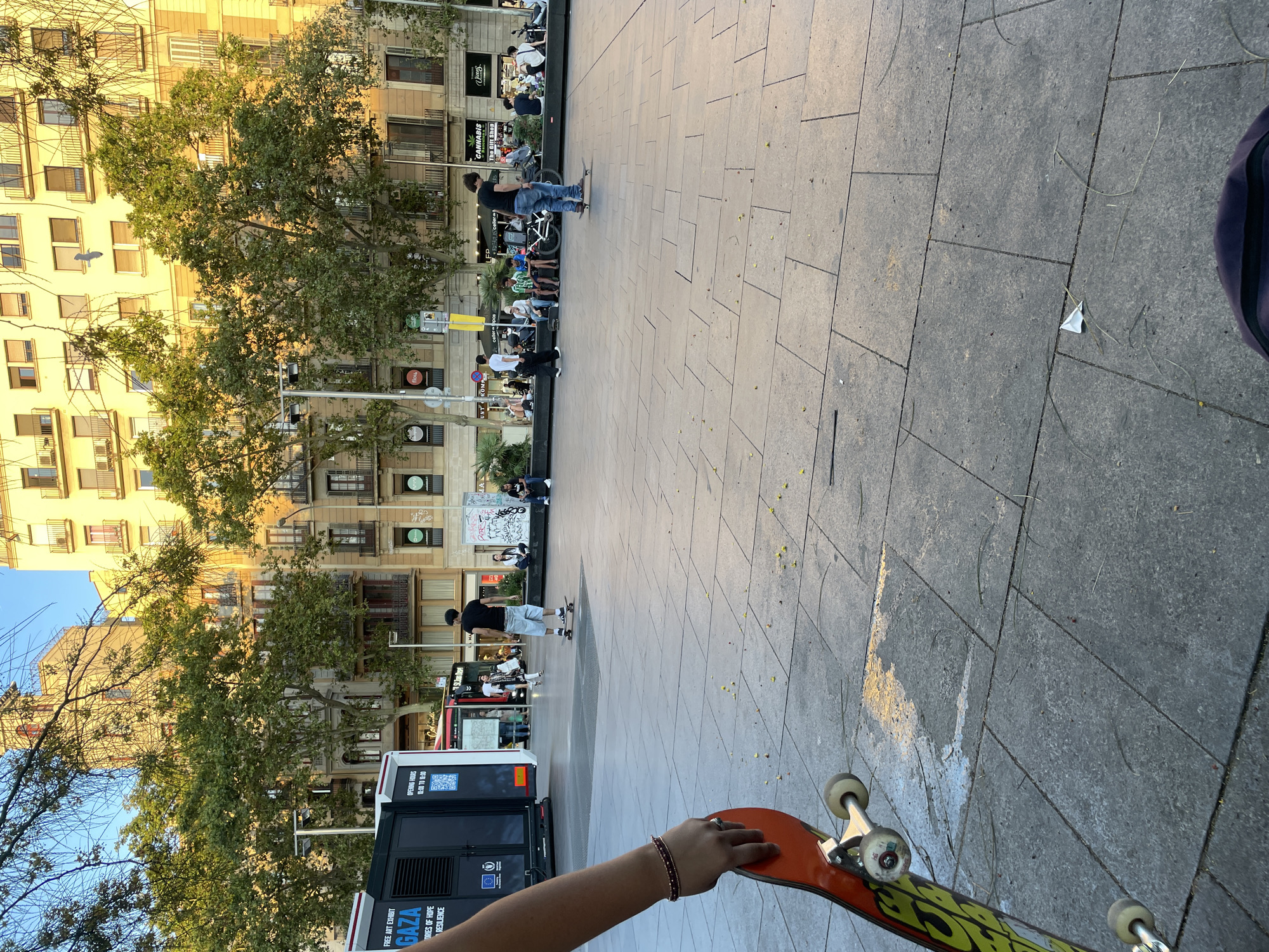 The image shows a public plaza with a group of people. In the foreground, a person's arm and hand are holding an orange and yellow skateboard. The plaza is paved with large gray tiles. In the mid-ground, several people are skateboarding, and others are sitting on benches or a low wall. One person is wearing a black t-shirt and blue shorts and is standing on a skateboard. Another person is wearing a black t-shirt and jeans and is also on a skateboard. In the background, there is a large building with multiple stories and balconies, and several trees with green foliage line the street. A black truck or trailer with text and graphics is parked on the left side of the plaza. The lighting suggests it is late afternoon or early evening.