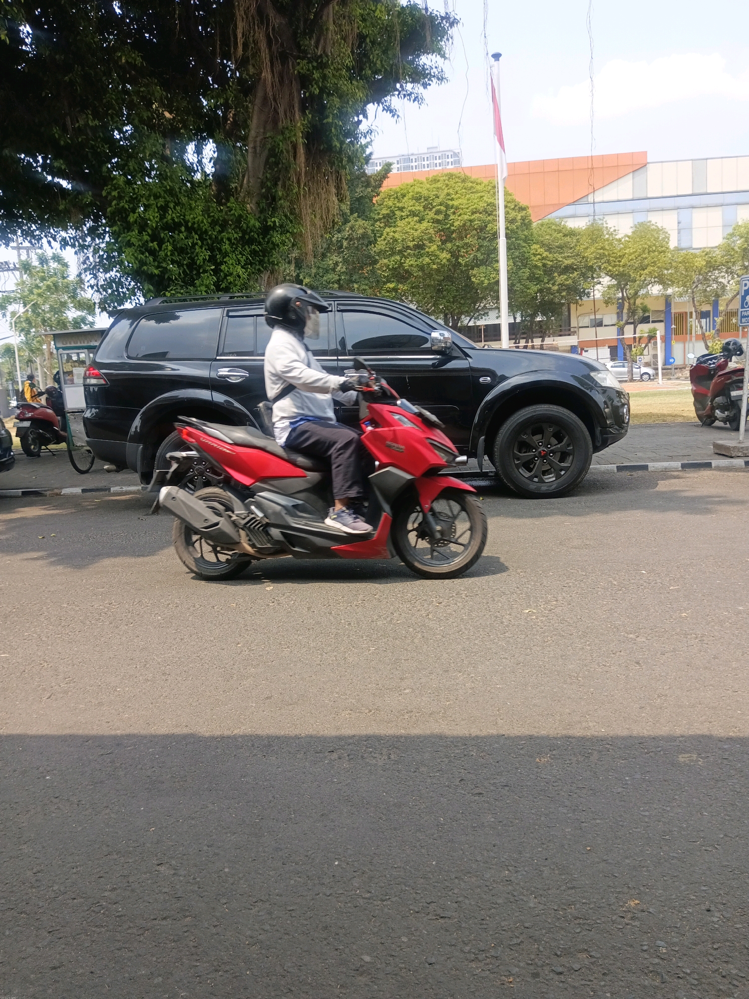 In Surabaya, Indonesia, a man wearing a black helmet and a light-colored long-sleeved shirt is riding a red motorcycle. He is wearing dark pants and light-colored sneakers. The motorcycle has black accents and silver wheels. To the left of the motorcycle, a black SUV is parked. The SUV has a black roof rack and chrome door handles. In the background, a large tree with hanging roots is visible, along with a building with orange and blue sections. A flag pole with a red and white flag is also present. The scene is on a paved road, likely a street or parking lot, with sunlight casting shadows. The area appears to be urban with greenery and modern architecture.