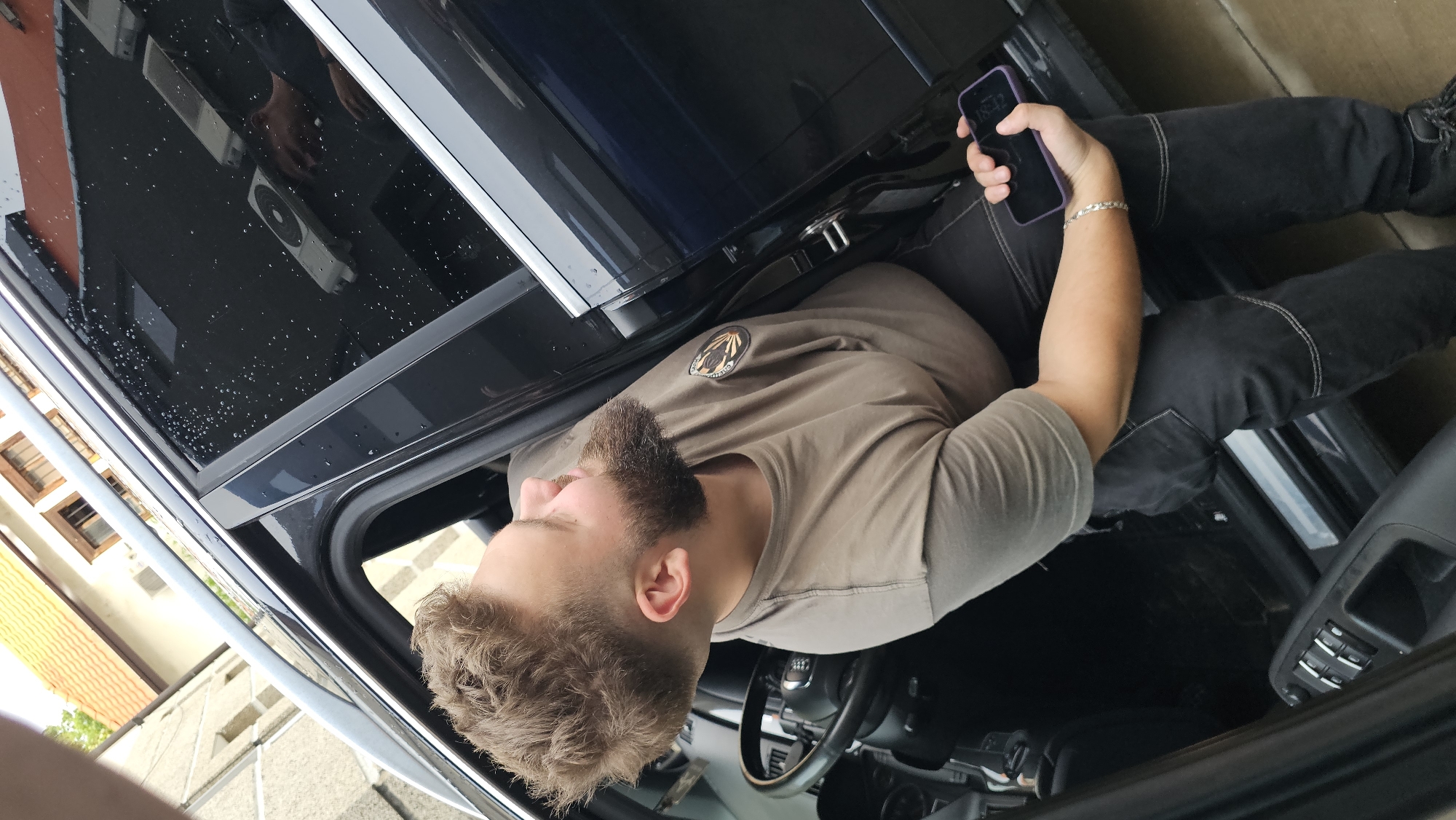 A Caucasian male with a beard and short, styled blonde hair is partially seated in the driver's side of a dark-colored SUV. He is wearing a tan t-shirt with a circular emblem on the left chest and dark jeans with reinforced stitching. His left arm rests on the steering wheel, and his right hand is holding a purple smartphone. The car door is open, revealing the interior which includes the steering wheel, dashboard, and window controls on the door panel. The window is wet with visible water droplets and reflects an external scene that includes a white air conditioning unit and a partial reflection of the man himself. The vehicle appears to be parked outdoors, possibly in a driveway or street, with residential buildings and a tiled roof visible in the background. The scene is set in Karlovo, Bulgaria.