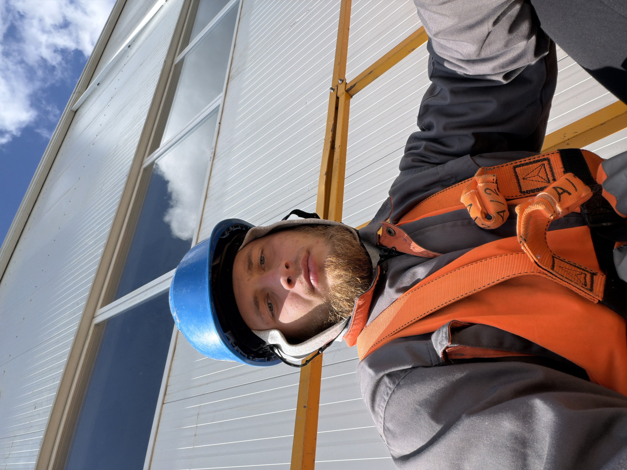 A medium shot captures a young man, likely in his early to mid-twenties, with a light complexion and a prominent ginger beard, looking directly at the camera. He is wearing a blue hard hat, a grey work jumpsuit, and an orange safety harness. The man appears to be outdoors, with a white, corrugated metal building and a clear blue sky with white clouds forming the background. A yellow metal structure, possibly scaffolding or part of the building's support system, is visible in the foreground, partially obscuring the man's torso and right arm. The lighting suggests it is daytime, with sunlight casting shadows on the man's face and clothing. The man's expression is neutral. The scene is devoid of any specific indicators of the city of Stróża, Poland, or any other urban context. The image focuses on the man and his work attire, with the building and sky providing a generic outdoor setting.