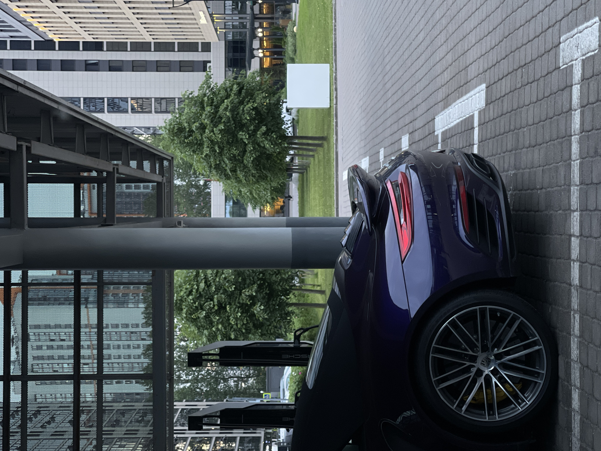 A dark purple sports car is parked in a designated parking spot on a paved surface. The car's rear wheel and a portion of its body are visible. In the background, several modern buildings with many windows are present. A row of trees lines a grassy area between the parking lot and the buildings. Two tall, dark charging stations are positioned near the car. The scene is outdoors, likely in an urban environment, under an overhead structure supported by large pillars.