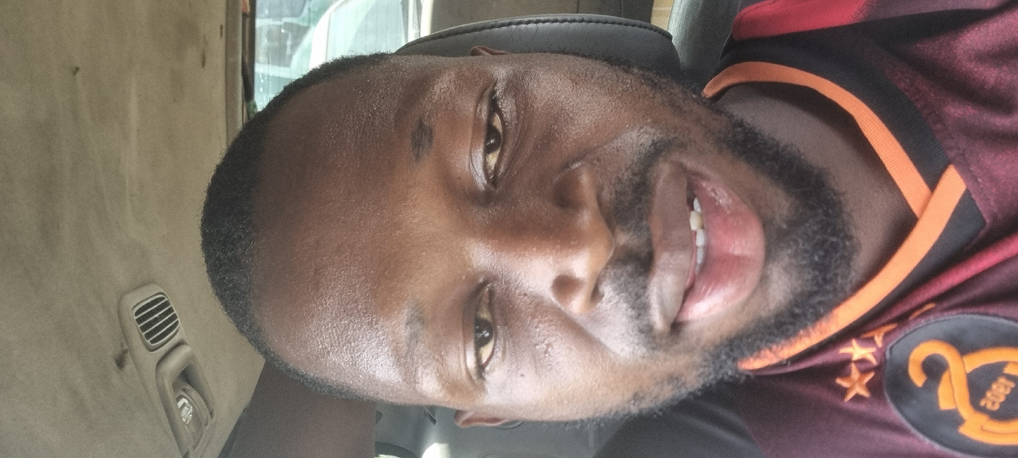 The image is a close-up selfie of a Black man with short black hair and a trimmed beard. He is wearing a maroon and orange soccer jersey featuring three stars and the text "2002". The man is inside a vehicle, likely a car or truck, as evidenced by the vehicle interior, including air vents, visible in the background. Based on the image information, it's impossible to confirm the location in Satellite Town, Nigeria. There are no other people, subjects, interactions, or environments visible.