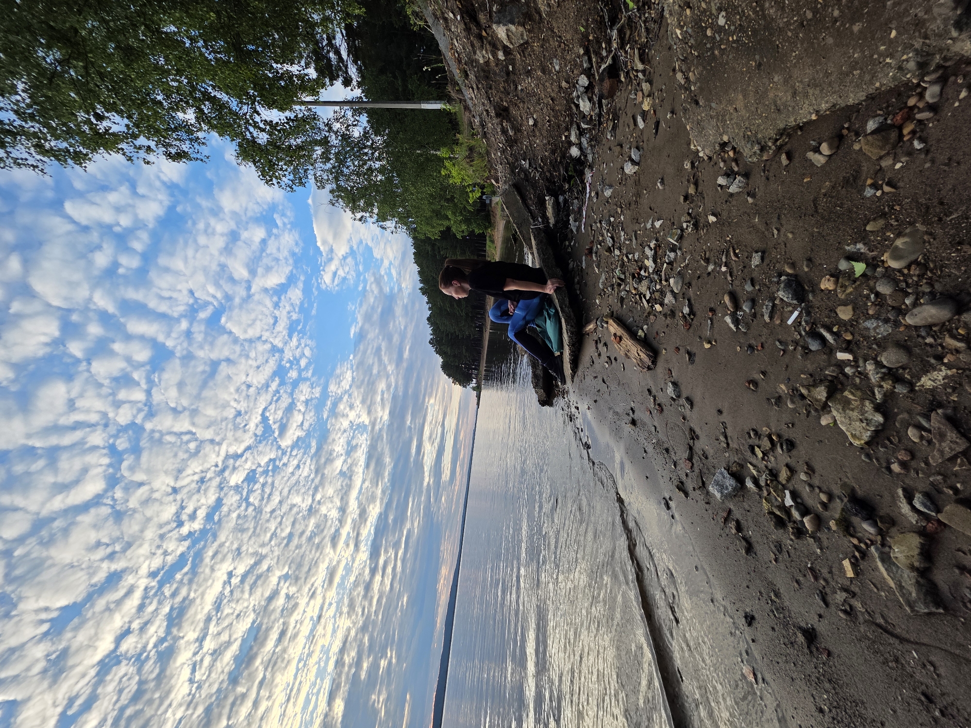 A young woman with long brown hair tied in a ponytail is sitting on a concrete slab on a rocky, sandy shore of a large body of water, likely a reservoir, based on the context of the city of Chaikovsky, Russia. She is wearing a black t-shirt and blue jeans. She is facing away from the camera, looking out towards the water and the horizon. A green backpack is placed on the ground next to her. The water is calm with gentle ripples. The shore is littered with rocks, pebbles, and some debris, including a piece of wood. In the background, there is a dense forest line of pine trees and deciduous trees. The sky is blue with scattered white, fluffy clouds, indicating it might be late afternoon or early evening, with the sun casting a soft light. A light pole is visible among the trees. The overall scene is one of natural beauty and quiet contemplation.
