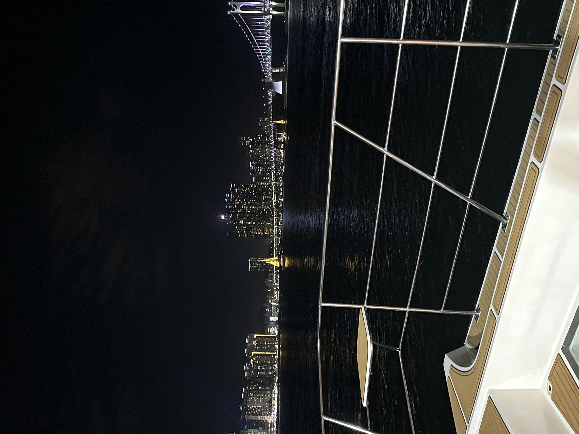 The image shows a nighttime view of the Gwangan Bridge and the Busan cityscape from the perspective of a boat on the water. The Gwangan Bridge, illuminated with colorful lights, is prominently featured on the right side of the frame, spanning across the dark water. The Busan skyline, with numerous high-rise buildings lit up, is visible in the background. In the middle of the water, a small, illuminated sailboat with a yellow sail is present. The foreground of the image displays a portion of the boat from which the photo is taken, including its railing and seating area, which has teak-colored decking. The water is dark and reflects the city lights. The sky is dark with some subtle cloud cover.