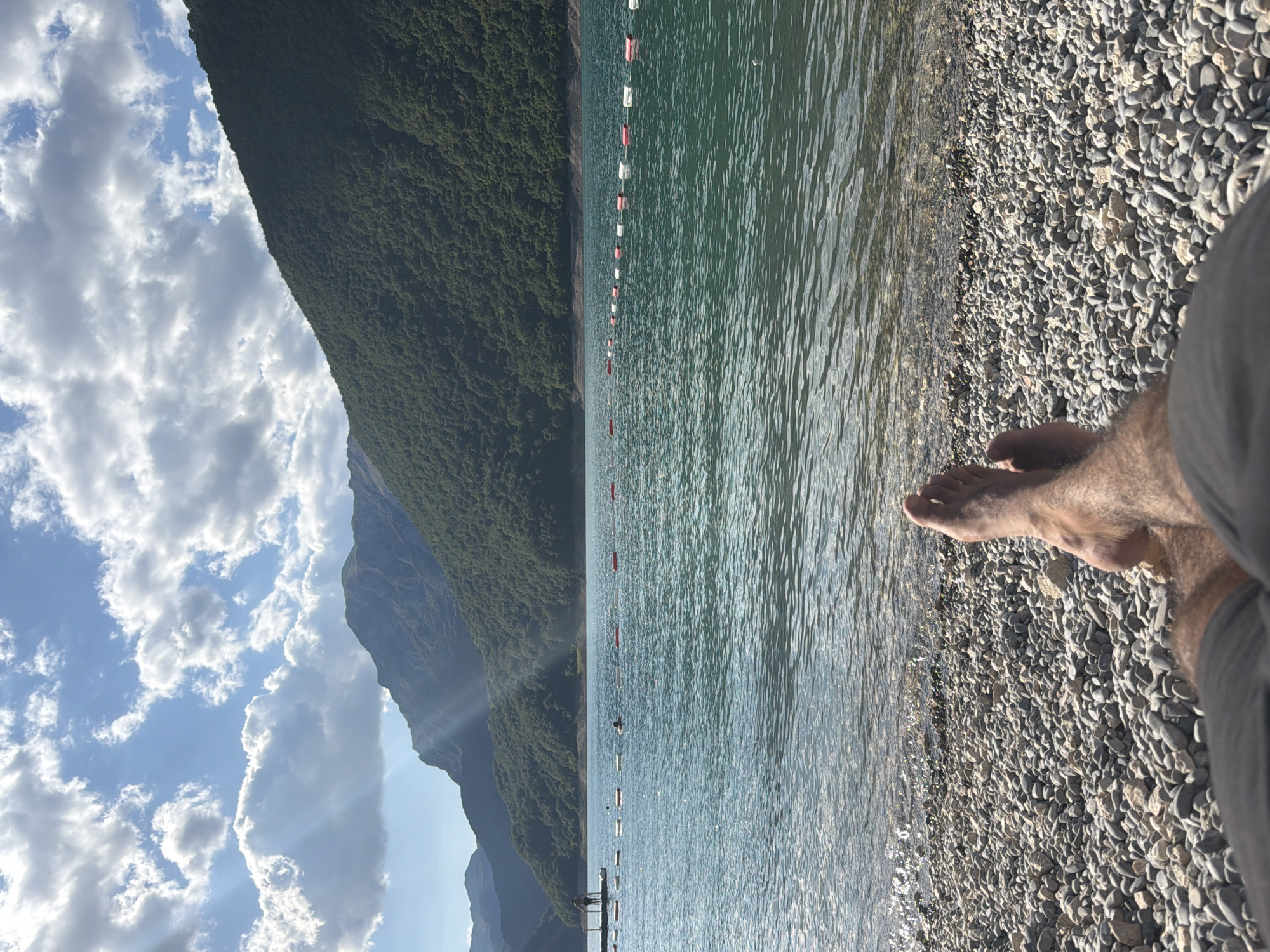 A first-person perspective shot shows legs and feet resting on a pebble beach, facing a wide, calm lake. The water is a deep turquoise and appears clear, with sunlight glinting off its surface. A line of buoys, alternating red and white, stretches across the water, demarcating a swimming area. In the distance, a lone figure stands on a dock. Lush, green, tree-covered mountains rise steeply on the far side of the lake, their peaks partially obscured by dramatic, sunlit clouds in a bright blue sky. The scene conveys a sense of peaceful relaxation in a natural, mountainous setting, likely the Piva Lake near Plužine, Montenegro, known for its stunning scenery.