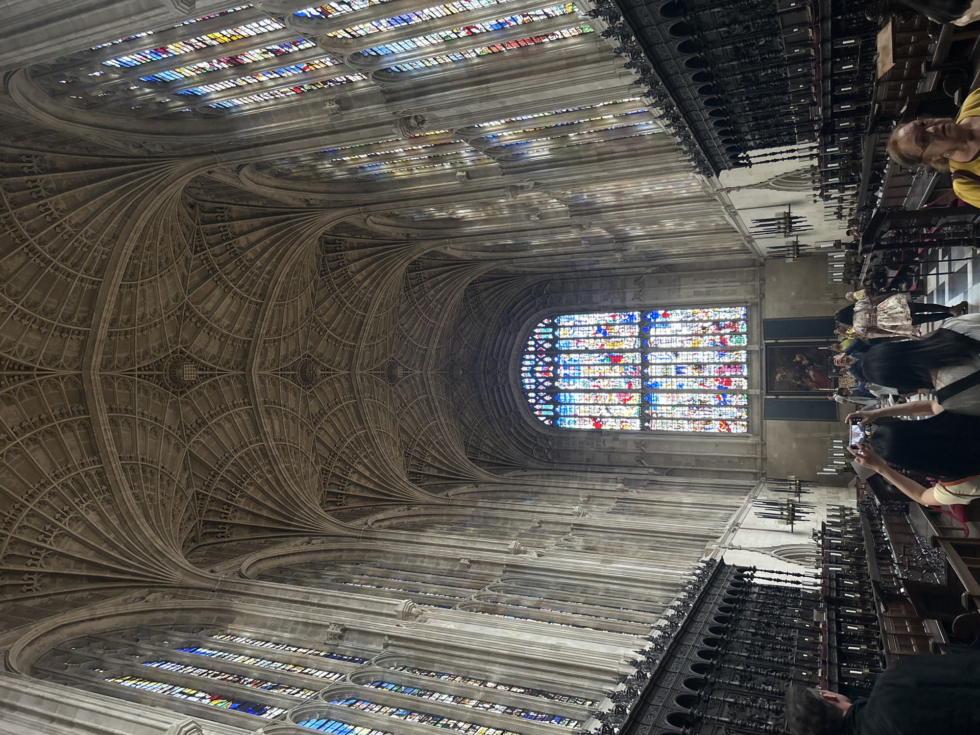 The image depicts the interior of King's College Chapel in Cambridge, United Kingdom. The chapel is characterized by its impressive fan vaulting ceiling and numerous stained-glass windows, which cast colorful light into the space. Long rows of dark wooden choir stalls line the sides of the chapel. Several people are visible, some walking, and others taking photographs. In the foreground, a person wearing a black jacket is seen from behind, facing towards the chapel's interior. To their right, two individuals are photographing the scene with their phones. Further down, a woman in a light-colored dress walks away from the camera, and another woman in a yellow top is looking towards the right of the frame. The environment is grand and architectural, with a sense of historical significance.