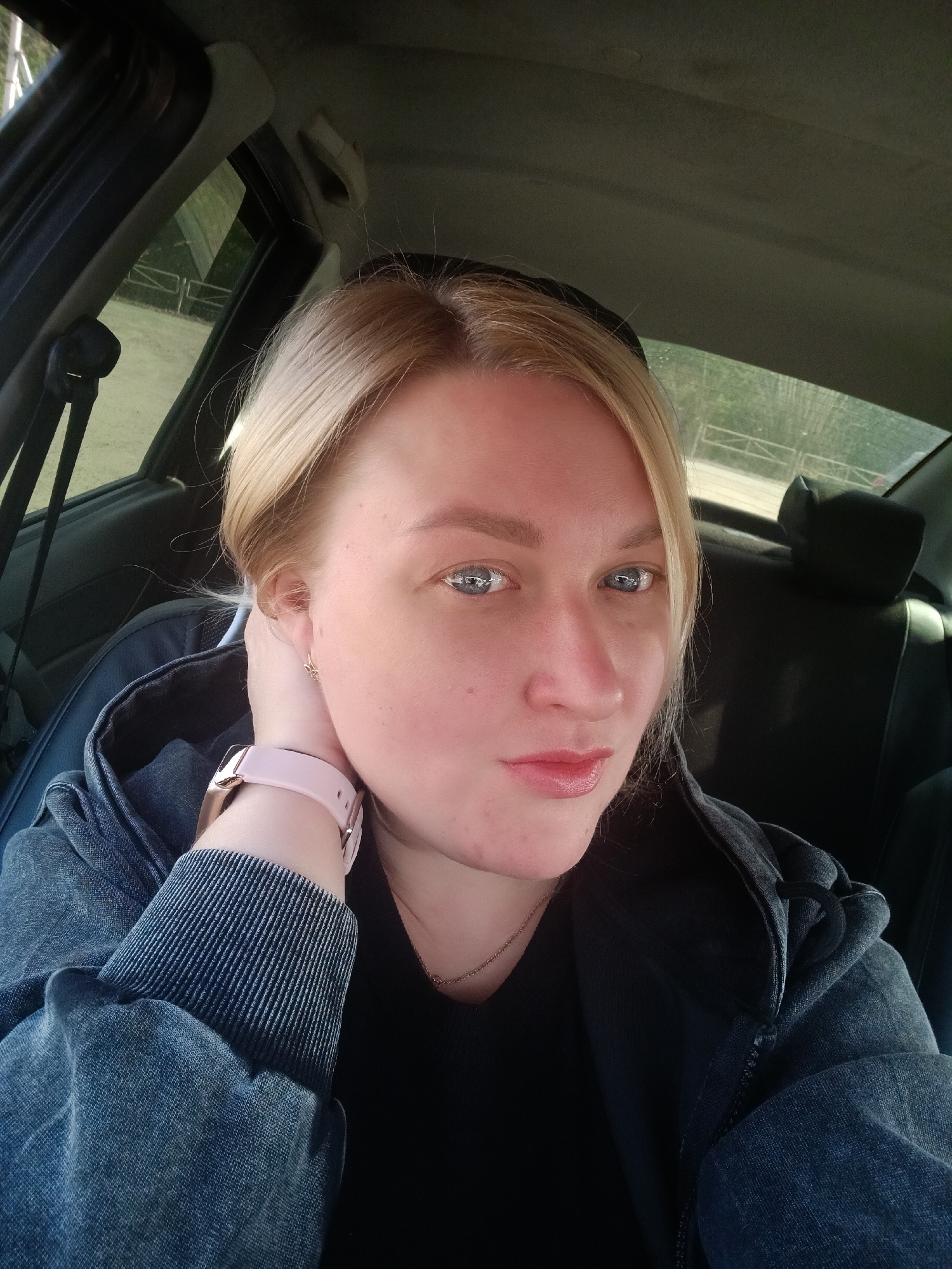 A selfie captures a woman seated inside a car, looking directly at the camera. She has blonde hair pulled back, light blue eyes, and fair skin with a slight flush. Her lips are a soft pink shade, and she wears subtle gold stud earrings. Her left hand is raised to her neck, revealing a light pink smartwatch with a rose gold buckle on her wrist. She is dressed in a black t-shirt beneath a dark denim jacket or hoodie.
The interior of the car is visible around her, with dark upholstery and parts of the car's frame. Through the car windows, a blurred outdoor scene is visible. On the left side, tall trees and what appears to be a light-colored fence or railing can be seen, suggesting a natural or semi-urban environment. The background on the right side also shows greenery and some distant structures or fences. The lighting suggests it is daytime, with sunlight illuminating the woman's face and creating reflections in her eyes. The overall mood is casual. There is no visible text in the image. The location is contextualized as Chaikovsky, Russia.