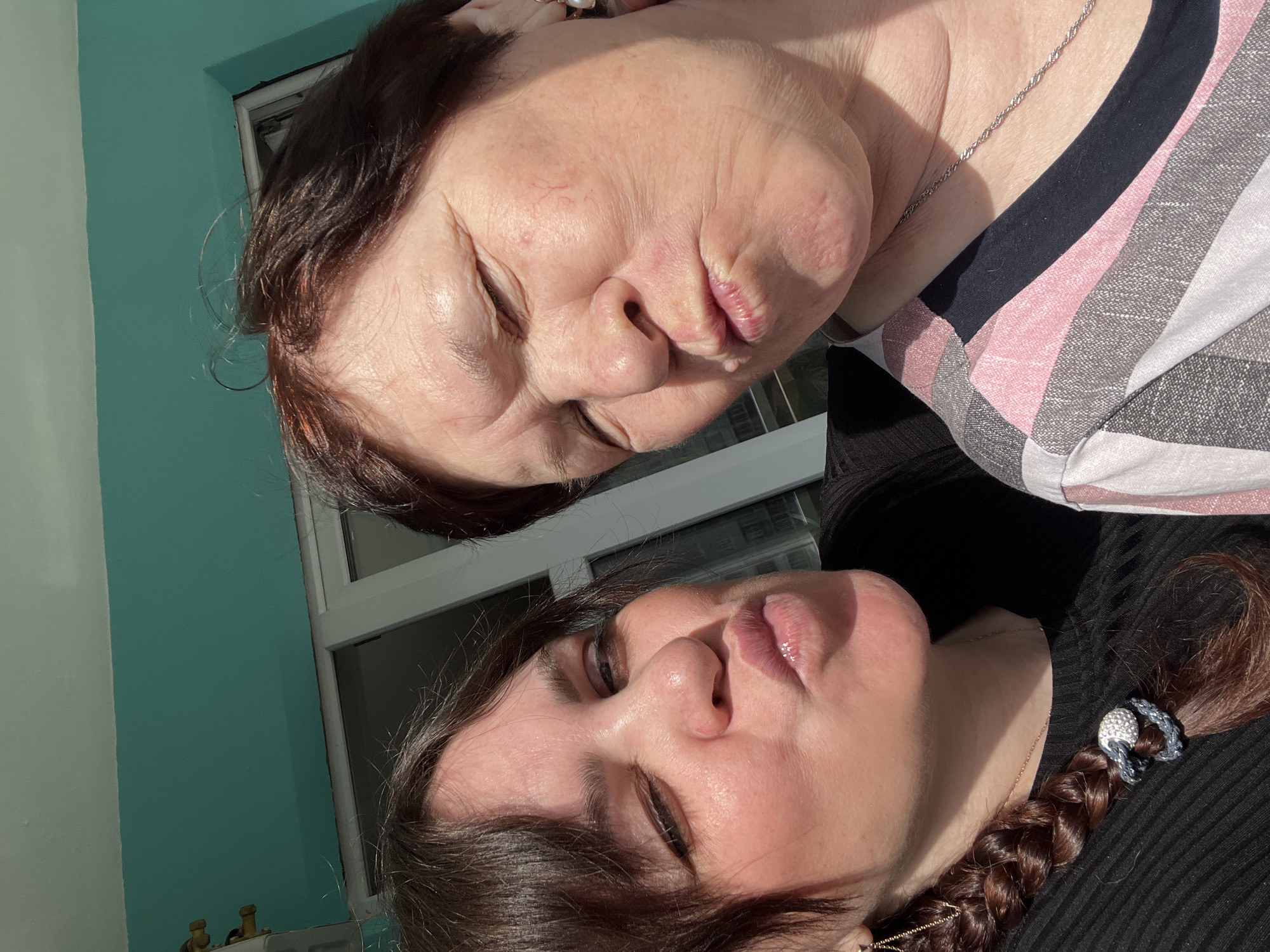 The image is a close-up selfie featuring two women, likely a mother and daughter, both posing with playful, pursed lips. They are positioned close together and tilted on their sides, suggesting a hand-held shot.
The woman at the top of the frame, appearing older, has dark brown hair and fair skin with visible wrinkles. She is wearing a striped top with patterns in pink, white, grey, and black, and a delicate silver chain necklace is visible around her neck.
The woman at the bottom of the frame, appearing younger, also has dark brown hair, styled in a braid secured with a silver/grey hair tie featuring sparkling details. She is wearing a dark, ribbed top, and a thin gold chain necklace is visible.
Both subjects are illuminated by bright, direct sunlight, which casts distinct shadows and highlights their features. The background includes a prominent teal or turquoise colored wall next to a white-framed window, through which bright light enters. Another lighter, possibly off-white, wall is partially visible. The strong sunlight indicates it is daytime. The overall mood conveyed is affectionate and playful. No text is visible in the image. The setting appears to be indoors, in Perm, Russia.