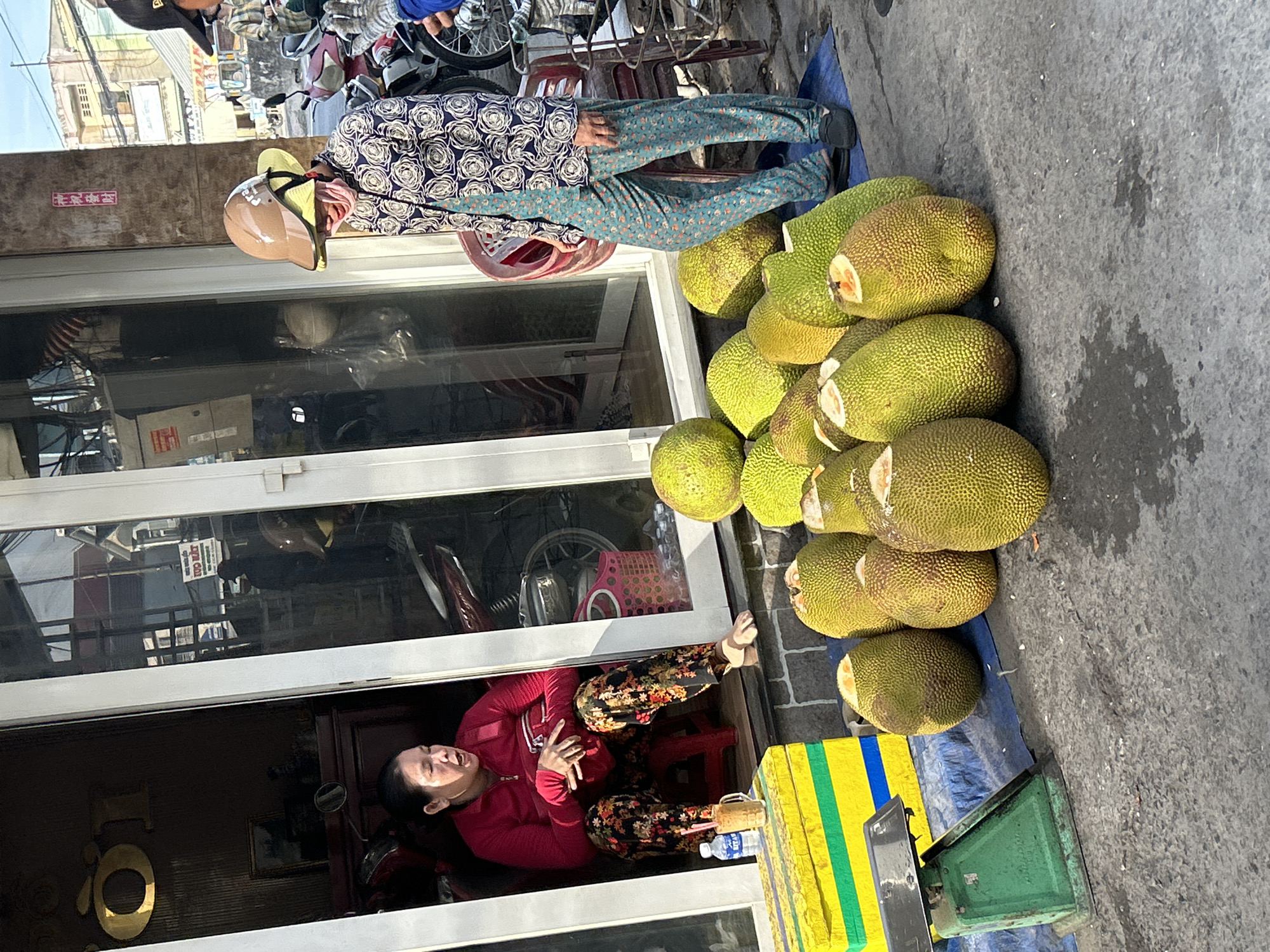 The image captures an outdoor market scene in An Hữu, Vietnam. In the foreground, a pile of ripe jackfruits are displayed on a blue tarp. To the left, a vintage green scale sits next to a yellow and green striped box. A woman in a red shirt and floral pants sits on a red stool inside a shop entrance, her mouth open as if speaking. To her right, a glass of iced beverage with a straw is visible. In the middle ground, a person wearing a patterned shirt, floral pants, and a helmet stands amidst the jackfruits, looking down. Behind them, a stack of red chairs and motorcycles are partially visible. The background shows buildings and typical street elements of a Vietnamese town.
