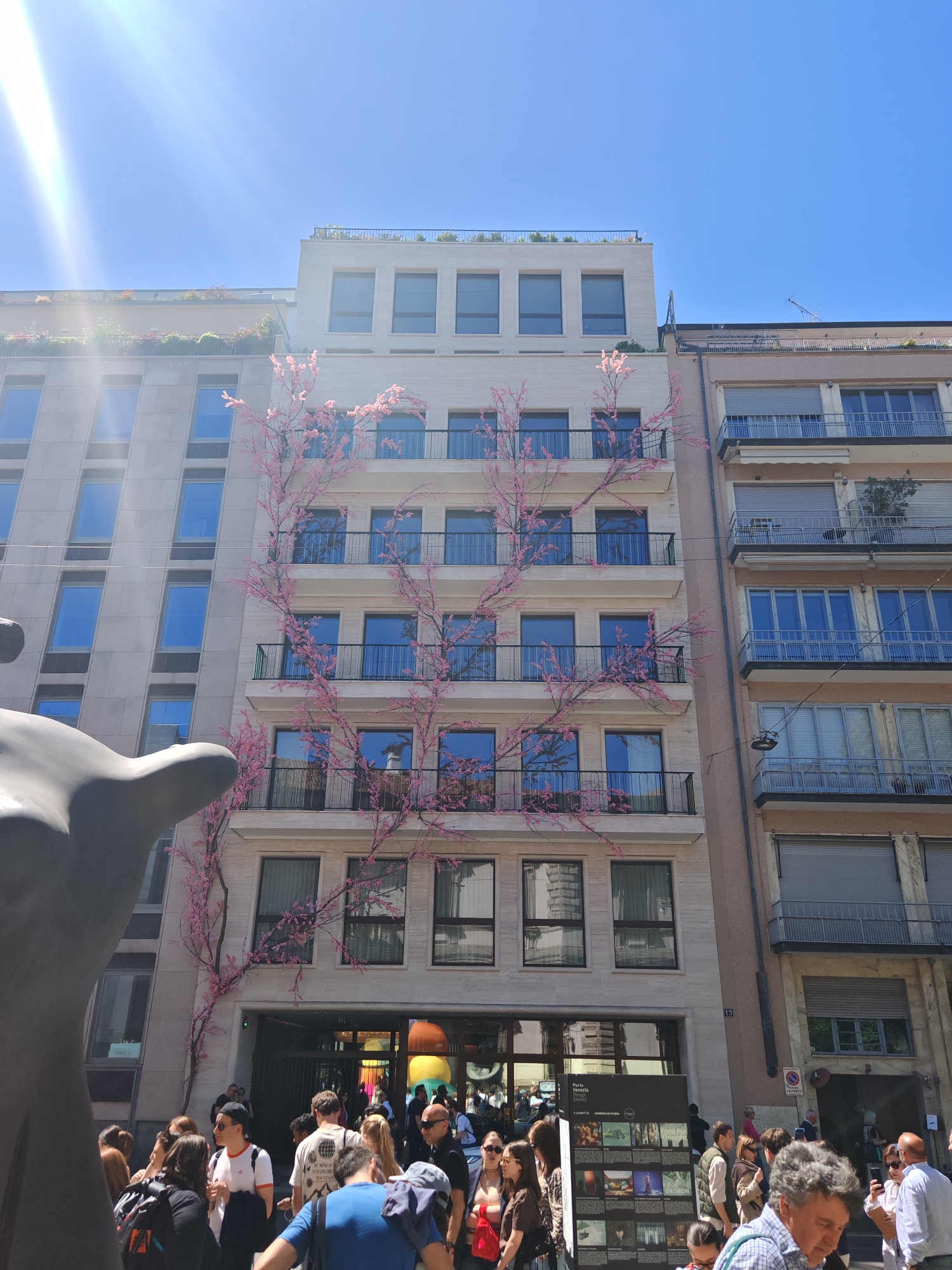The photograph captures a bustling urban scene. Several buildings line the street, including one adorned with pink flowering branches that climb several stories. This building features numerous windows and balconies. People are gathered near the entrance of a building. In the foreground, there appears to be a statue on the left, partially cropped, and a poster display. The weather seems sunny and clear.