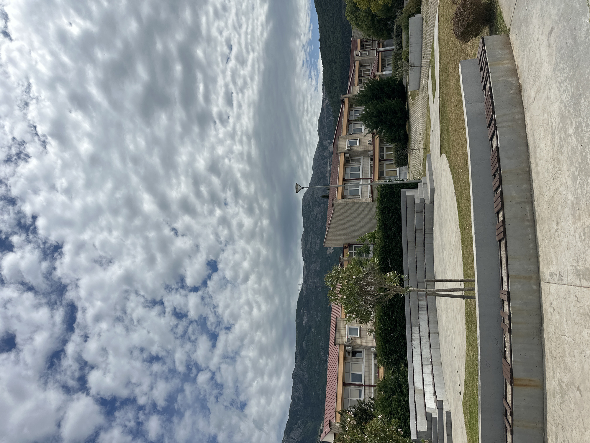 The image captures a serene urban landscape against a dramatic natural backdrop, likely in Budva, Montenegro, under a captivating sky.
The upper portion of the scene is dominated by a vast, dynamic sky filled with a mixture of dense and scattered altocumulus clouds. Patches of clear blue sky are visible, particularly towards the left, contrasting with the predominantly grey and white cloud cover which forms intriguing, wave-like patterns across the expanse.
In the background, a rugged and imposing mountain range stretches across the horizon. These mountains are heavily covered in dense green vegetation, indicating a forested and natural landscape that rises steeply behind the urban development.
The midground features a multi-story building complex, possibly apartments or a hotel. The buildings exhibit light-colored facades, which appear to be constructed from small tiles or stucco, and are topped with distinct reddish-brown or terracotta-colored roofs. Numerous windows are visible, many with external air conditioning units mounted beneath them. Lush green hedges and trees are planted along the front and between the buildings. A tall, slender street lamp stands in front of one of the structures.
The foreground presents a modern, landscaped public area. It consists of broad concrete pathways and terraced steps interspersed with neatly maintained grassy sections. A prominent feature on the right is a contemporary bench, crafted from concrete supports with wooden slats, designed for public seating. Several small, young trees with slender trunks are planted within the grassy areas, contributing to the green aesthetic of the structured environment.
No people or specific activities are visible in the image, conveying a sense of calm and stillness in this public space. The time of day appears to be daytime, likely late morning or early afternoon, under bright but diffused light from the dramatic, cloud-filled sky. The architecture, mountainous terrain, and public area design are consistent with the provided location context of Budva, Montenegro. There is no visible text in the media file.