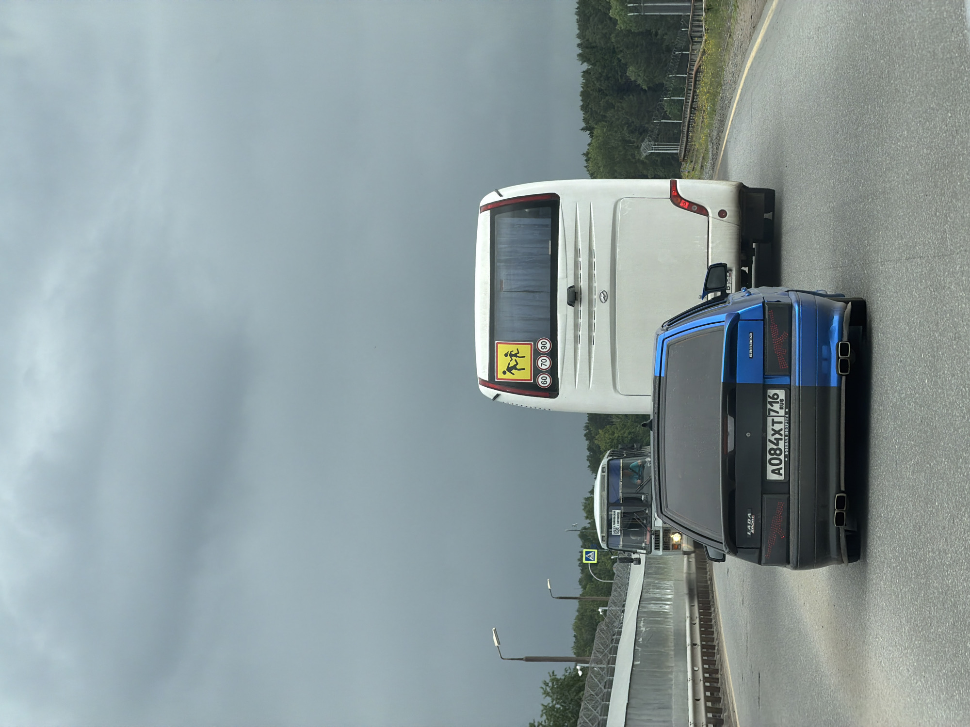 The image captures a three-vehicle traffic scene on an asphalt road under a heavily overcast, dark grey sky. In the foreground, a modified Lada Samara sedan is visible from the rear, distinguished by a unique black and blue split-color paint scheme (left half black, right half blue). The car displays a Russian license plate, A084XT 716 RUS, with the text "БОЕВАЯ ПОДРУГА" (Combat Girlfriend) in Cyrillic below it. It features custom aftermarket taillights with a red matrix pattern and dual exhaust tips on both the left and right sides.
Directly in front of the Lada is a white passenger coach bus, seen from its rear. The bus has a yellow square sign affixed to its back, depicting two black stick figures (children) and speed limit numbers "60", "70", and "90" (partially visible), indicating it is a children's transport vehicle.
Further down the road, in the same lane as the coach bus, an older white municipal bus is visible. Its front destination sign clearly displays "РУСЬ ЧАЙКОВСКОГО БОЛОТОНОВО" in Cyrillic, identifying the location as Chaikovsky, Russia. Above this bus, a yellow square sign with a black exclamation mark (general warning) and another yellow sign depicting a pedestrian crossing are visible.
To the left of the road, a concrete barrier topped with razor wire extends along the roadside, accompanied by streetlights. Dense green foliage and trees line both sides of the road in the midground and background. The asphalt road appears damp or recently wet, with a continuous yellow line marking the right shoulder.