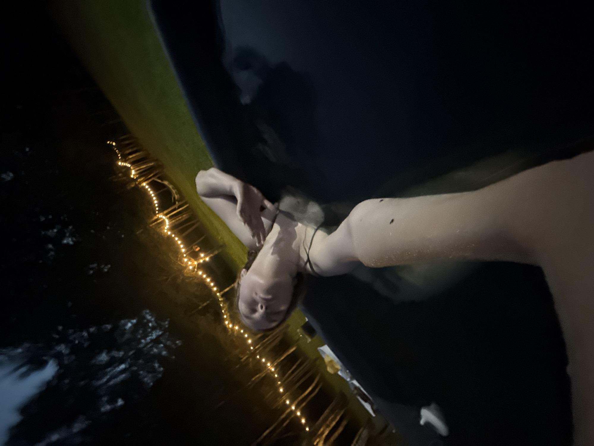 An outdoor, eye-level, slightly tilted selfie captures a light-skinned individual partially submerged in dark water within a black tub at night in the city of Копнино, Russia. The person, positioned centrally, has their head tilted back with eyes seemingly closed, displaying a relaxed demeanor. Their dark, wet hair is visible around their face. The left arm (from the subject's perspective) is bent, while the right arm extends toward the camera, likely holding the device. Small, reflective water droplets are visible on the skin. To the left of the tub, on a visible surface, a dark beverage in a glass and several light-colored food items are present. The background, illuminated by numerous warm-toned string lights strung horizontally across the scene, reveals the silhouettes of dark trees and a patch of green grass, indicating a nocturnal, natural setting. The sky is dark, consistent with nighttime.