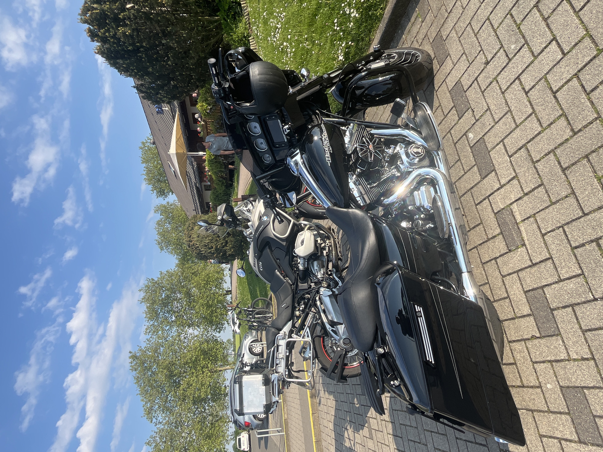 The image depicts a scene in Mönchengladbach, Germany, featuring several motorcycles parked on a brick-paved area. Two prominent black motorcycles, likely Harley-Davidson models, are in the foreground. To the left, further into the background, are other vehicles including a silver car and another motorcycle, possibly a BMW adventure bike with panniers. Beyond the motorcycles, some people are standing near a building with an awning, possibly a restaurant or cafe. The sky is blue with scattered clouds. The setting appears to be an outdoor parking area.