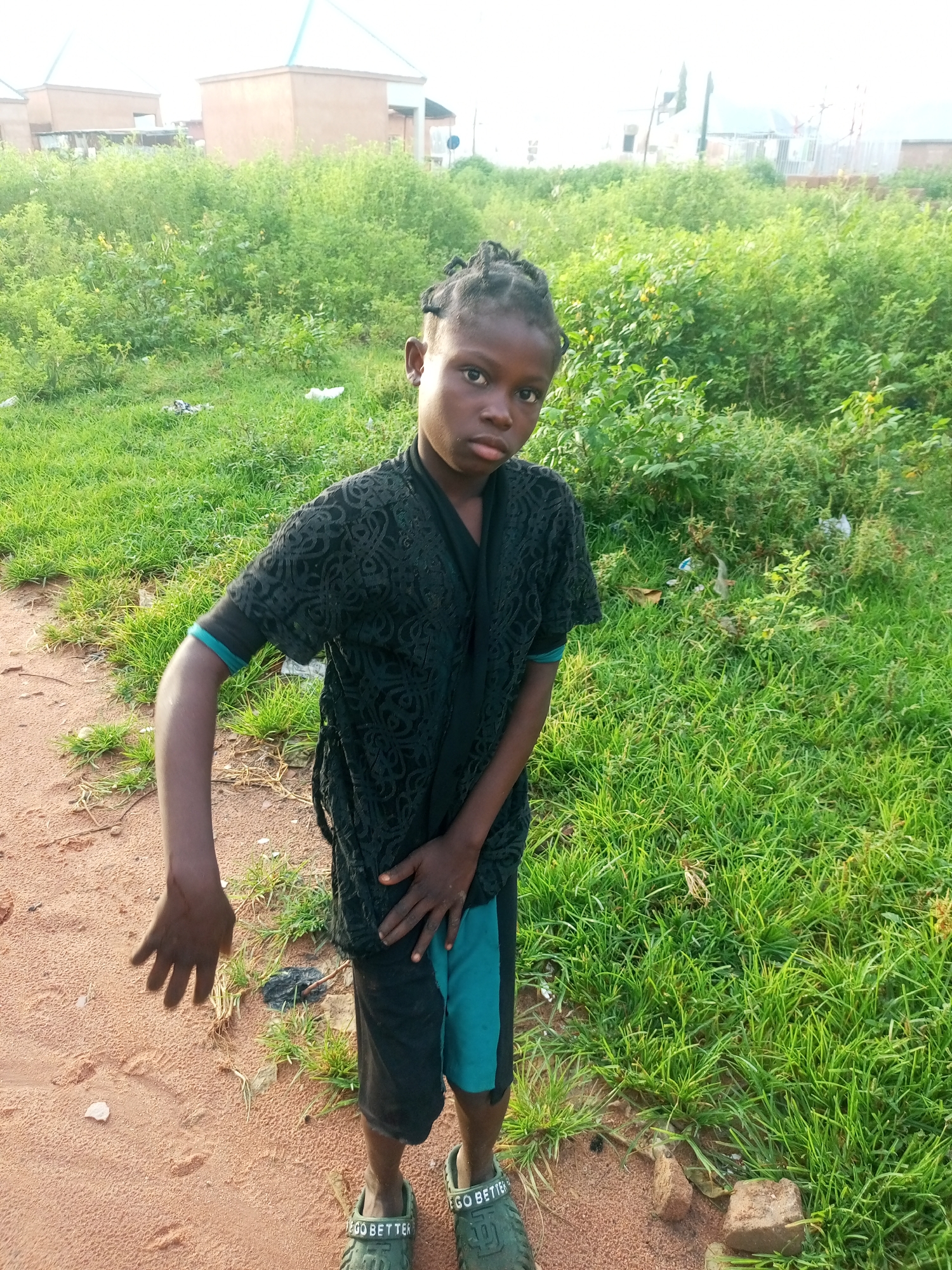 In Tempure, Nigeria, a young person stands on a sandy path beside a grassy area. They are wearing a black patterned shirt over a dark garment, black shorts with teal accents, and green Crocs-style sandals. Their left arm is bent and held forward, while their right hand rests on their shorts. The background features lush green vegetation, and several buildings are visible in the distance. The sky is bright, suggesting daytime.