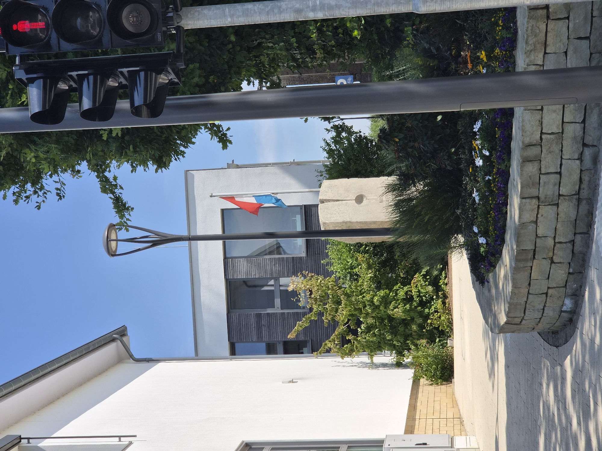 This image captures an outdoor scene on a clear, sunny day, likely in the afternoon due to the strong shadows cast on the pavement. The composition is oriented vertically.
In the foreground, a modern white building with a gray-tiled roof is visible on the left. To the right of the building, a pathway paved with light-colored stones leads towards a landscaped area. This area features a low stone wall constructed from irregular blocks, and a variety of plants, including green bushes and flowering plants with white and purple blossoms.
In the middle ground, a modern building with white upper walls and a dark wooden-paneled section is prominent. A flagpole with the flag of Luxembourg (red, white, and light blue horizontal stripes) is mounted in front of this building. Adjacent to the flagpole is a rough, stone monument or marker with etched lines and a circular indentation, suggesting it might be a sundial or a boundary marker. A streetlamp with a distinctive modern design stands near this monument.
In the background, more lush green trees and a bright blue sky are visible. A traffic signal with three black housings is mounted on a metal pole at the top left of the frame. The red arrow light is illuminated, indicating traffic direction.
The overall impression is one of a well-maintained public space, possibly in a town or village in Luxembourg, given the flag and the presence of a traffic signal. The scene evokes a sense of calm and pleasant weather. There is no visible text other than on the traffic signal, which reads "TRAFFIC".