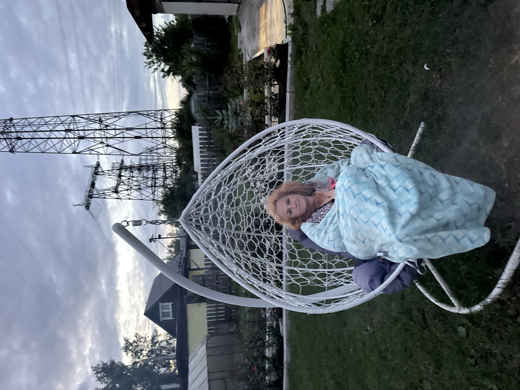 The image captures an outdoor scene in the city of Chaikovsky, Russia, characterized by a low-light ambiance, possibly dusk. In the foreground, a woman is seated in a white, egg-shaped hanging chair, which is suspended by a metal stand. She is wrapped in a light blue blanket with a diamond pattern and appears to be holding a glass in her right hand. Her hair is a vibrant shade of red.
The background reveals a residential area with houses, a greenhouse, and various trees and shrubs. Dominating the upper portion of the image are tall, lattice-like electrical transmission towers and power lines, indicative of the industrial infrastructure often found in or near Russian cities. The ground is covered with grass, and there are paved pathways. The sky is overcast with heavy clouds.