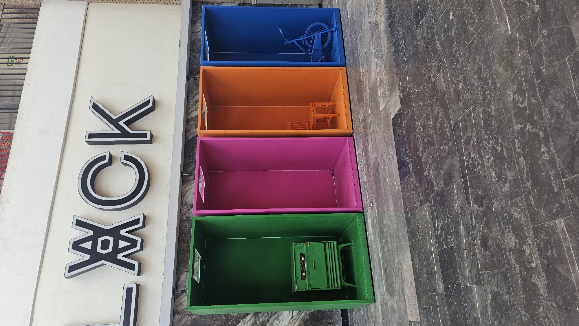 This eye-level photograph, taken in Puebla, Mexico, shows a stack of four brightly colored open-topped containers next to a store sign with the name "LOCK" vertically written. The background includes a gray stone sidewalk.
The containers are stacked vertically, one on top of the other. From top to bottom, they are blue, orange, pink, and green. Each container appears to hold a small object. Inside the blue container, there's a small toy bicycle. The orange container holds a miniature chair. The pink container is empty. The green container has a toy-like device inside.
To the left of the containers, there's a vertical store sign that reads "LOCK". The letters are in a white font with a black outline.
The photograph has a slight blur near the top right corner, but otherwise, it is in sharp focus. The lighting appears to be natural daylight.