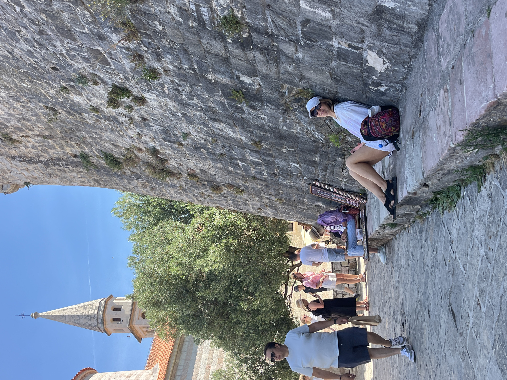 In Budva, Montenegro, on a sunny day, a street scene unfolds near an ancient stone wall. A man in a white t-shirt and dark shorts walks on a stone pathway, carrying a brown paper bag. Further down the path, a group of people are gathered, including a woman in a black dress and straw hat, and a man in a white t-shirt and light-colored shorts. To the right, leaning against the imposing stone wall, a woman in a white shirt, white cap, and sunglasses relaxes, with a colorful backpack beside her. In the foreground, an artist, wearing a straw hat and purple shirt, sits on a stone bench, appearing to draw or sketch. The background features a prominent bell tower with a spire, characteristic of Budva's historic architecture, and lush green trees. The stone wall displays visible signs of age and weathering, with small plants growing in its crevices.