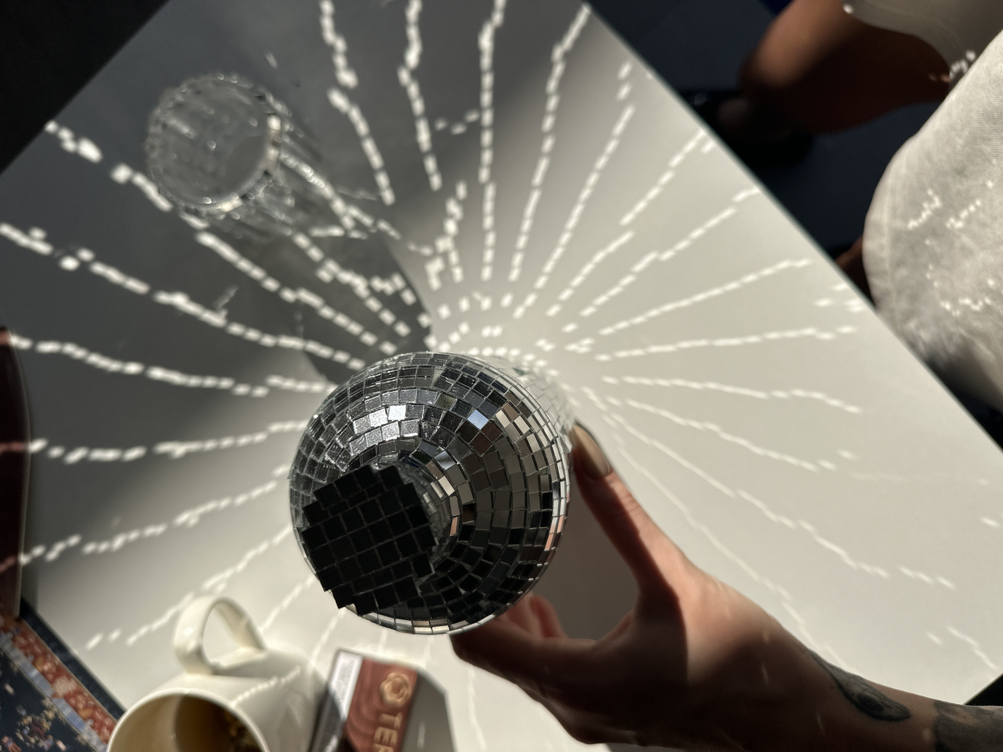 A hand holds a disco ball casting a pattern of light and shadow on a white surface. To the left, a white mug with a handle contains a light-colored liquid and a small tea bag tag is visible. In the upper right, a clear glass is positioned on the surface. The image appears to be taken indoors, with natural light streaming in, creating a dynamic interplay of light and shadow from what appears to be a nearby object with a patterned surface. The setting suggests a casual or domestic environment, possibly a table in a room. The overall aesthetic is bright and reflective. No specific location within North Kuta, Indonesia can be determined from the visual information provided.