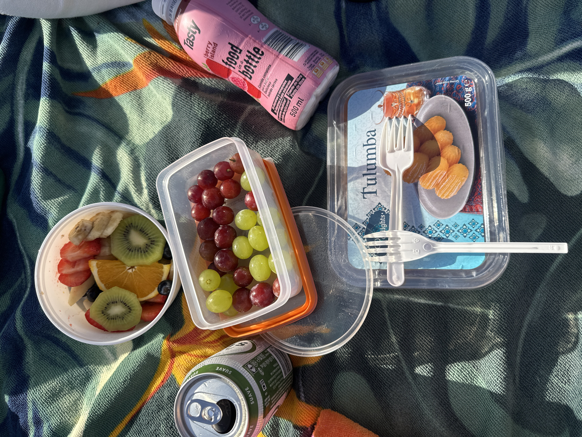 An overhead, eye-level shot captures a picnic spread on a patterned blue and green towel, illuminated by sunlight creating strong shadows. In a white bowl, a colorful assortment of fruit slices includes strawberries, kiwis, oranges, blueberries, and bananas. Adjacent is a clear plastic container holding red and green grapes. A silver can with a green label and the word "SUAVE" is positioned to the left. To the right, a pink bottle labeled "Tasty food in a bottle" and "Berry Island" stands upright. In the foreground, a clear plastic container showcases a serving of golden-brown, syrup-coated pastries labeled "Tulumba" with a 500g weight indication. A white plastic fork rests on top of the Tulumba container. The background is dominated by the textured pattern of the towel. The scene is consistent with a picnic in a public outdoor space.