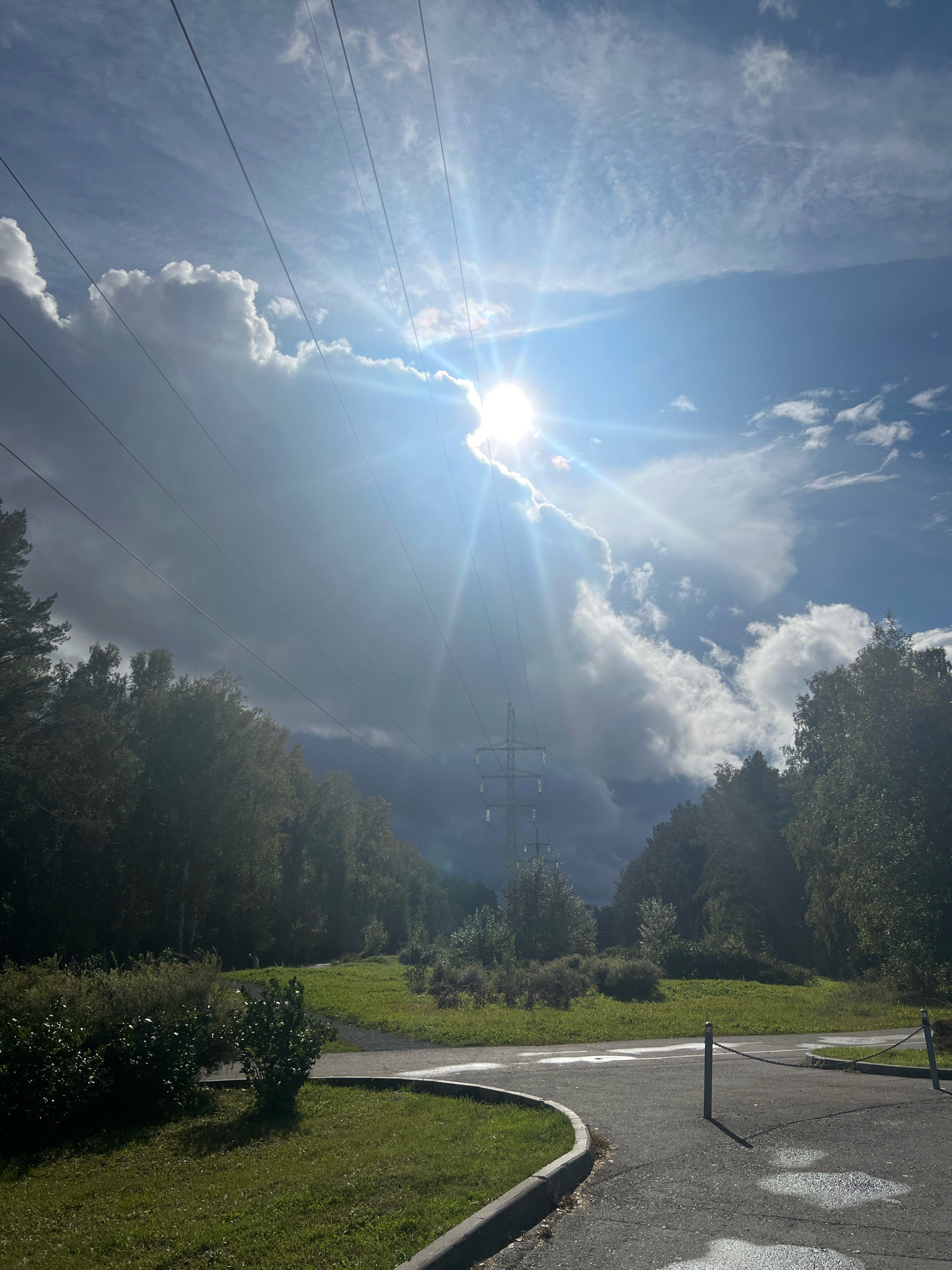 The image depicts a scene with power lines stretching across a bright sky, partially obscured by dramatic, sunlit clouds. The sun is a prominent source of light, creating a lens flare effect and casting rays across the frame. Below the sky, a landscape unfolds with a line of trees bordering a grassy area and a paved path. The path is wet in places, suggesting recent rain. In the distance, a tall, lattice-like transmission tower stands out. The overall impression is one of nature meeting infrastructure under a dynamic sky.
This image was taken at the Torgashinsky Ridge (also known as the Torgashinsky Trail or Torgashinskaya Step) in the city of Krasnoyarsk, Russia. The Torgashinsky Ridge is a popular nature reserve and recreational area located on the outskirts of the city, known for its walking trails and scenic views. The presence of the power lines and transmission tower is consistent with the infrastructure found in such natural areas that are accessible to urban populations. The trees visible are likely part of the taiga forest that covers the region.