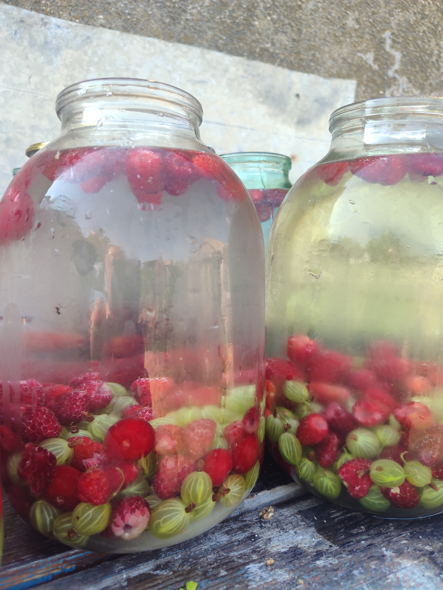 In Novosamarka, Ukraine, three large glass jars filled with raspberries and gooseberries submerged in liquid are placed on a weathered wooden surface. The jar on the left, largest of the three, is partially obscured by condensation and contains a dense collection of red raspberries and green gooseberries. A second, slightly smaller jar to its right contains a similar mix of berries and a clearer, yellowish liquid. Behind these, a smaller, greenish-tinted jar is visible, also containing berries. The background consists of a textured, grey surface, possibly concrete, with a hint of greenery suggesting an outdoor setting. The lighting indicates daytime.