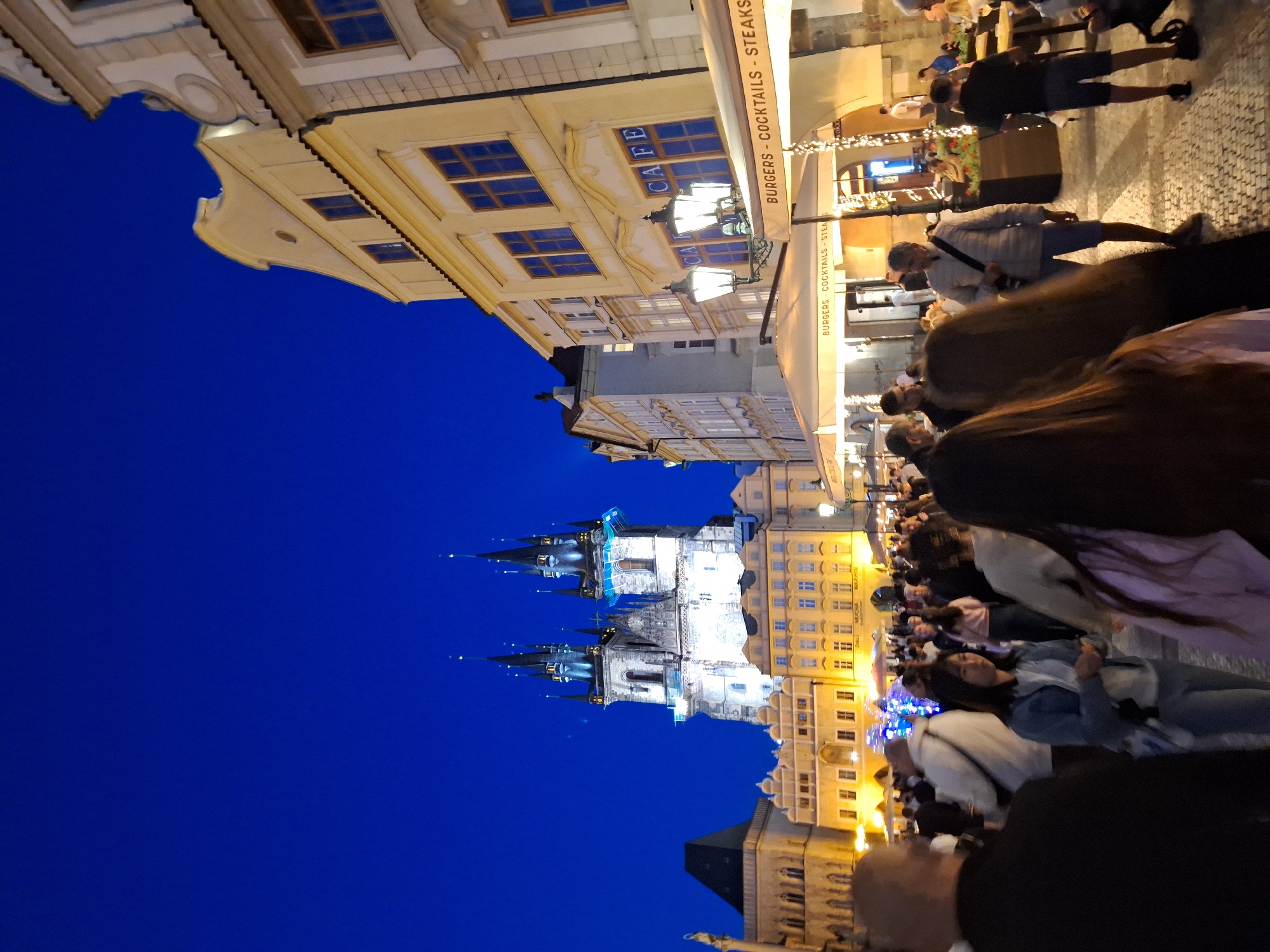 The image depicts a nighttime scene in Prague, Czechia, specifically in the Old Town Square. In the background, the iconic Týn Church with its Gothic spires is illuminated against a deep blue twilight sky. In the mid-ground and foreground, there are numerous people walking and standing, some gathered around outdoor seating areas of establishments. Buildings with traditional architecture line the square, some featuring lit windows and ornate facades. Several outdoor dining tents with signage reading "BURGERS - COCKTAILS - STEAKS" are visible, indicating a vibrant commercial area. Street lamps with ornate designs are also present. The overall atmosphere is one of a bustling urban gathering at dusk.