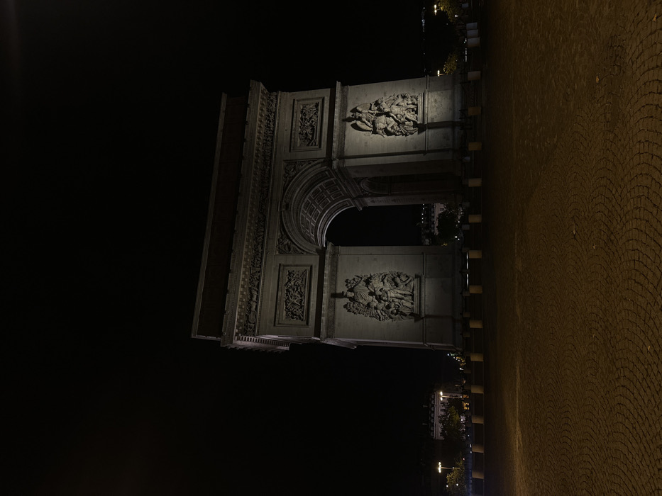 The image depicts the Arc de Triomphe at night, centrally located in Place Charles de Gaulle (Étoile), Paris, France. The monumental stone arch is brightly illuminated by artificial floodlights, highlighting its architectural details including the large sculptural reliefs on its facade, friezes, and cornices. The sky above is completely dark, indicative of nighttime. In the foreground, a wide expanse of cobblestone pavement, arranged in a repeating fan or scale pattern, appears empty and is illuminated with a warm, amber hue. A series of uniform, cylindrical, light-colored bollards forms a perimeter barrier between the paved foreground and the base of the monument. Faint outlines of distant buildings with scattered lights and shadowy trees are visible beyond the arch and to its sides. No individuals, vehicles, or observable actions are present within the scene.