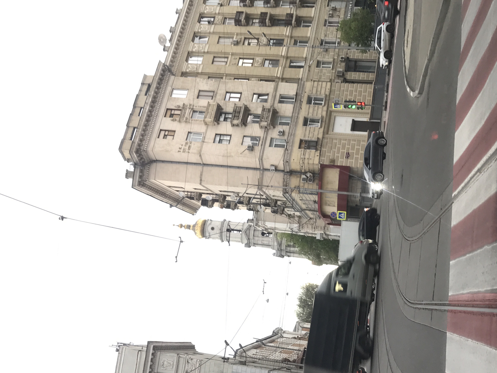 The image depicts a street scene in Kharkiv, Ukraine. A large, tan-colored building with multiple windows and balconies is visible on the left. Further down the street, a church with a golden dome can be seen. Power lines crisscross the sky. On the right, a street with a red and white striped curb is visible. Cars are driving on the road, including a black truck and a white car. A traffic light shows a red light. The sky is overcast.