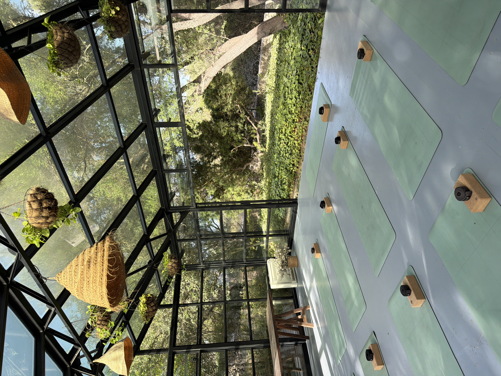This is a full shot of a yoga studio with a glass roof and walls, allowing for a view of the surrounding greenery. The studio has a grey floor, with multiple green yoga mats laid out, each equipped with two cork yoga blocks and a strap. In the background, there's a wooden table on trestle legs, a white heater, and a basket with dried flowers. The studio is filled with natural light, casting shadows on the floor. The view outside the glass walls shows trees, bushes, and a stone wall, suggesting a natural, possibly rural setting. The overall impression is one of tranquility and connection with nature. The yoga studio is located in Montuïri, Spain.